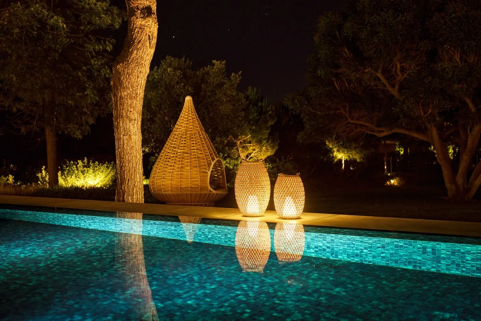 Nighttime scene of a swimming pool with illuminated mosaic tiles, surrounded by trees, with woven lanterns and a teardrop-shaped wicker chair on the poolside. Nestrest