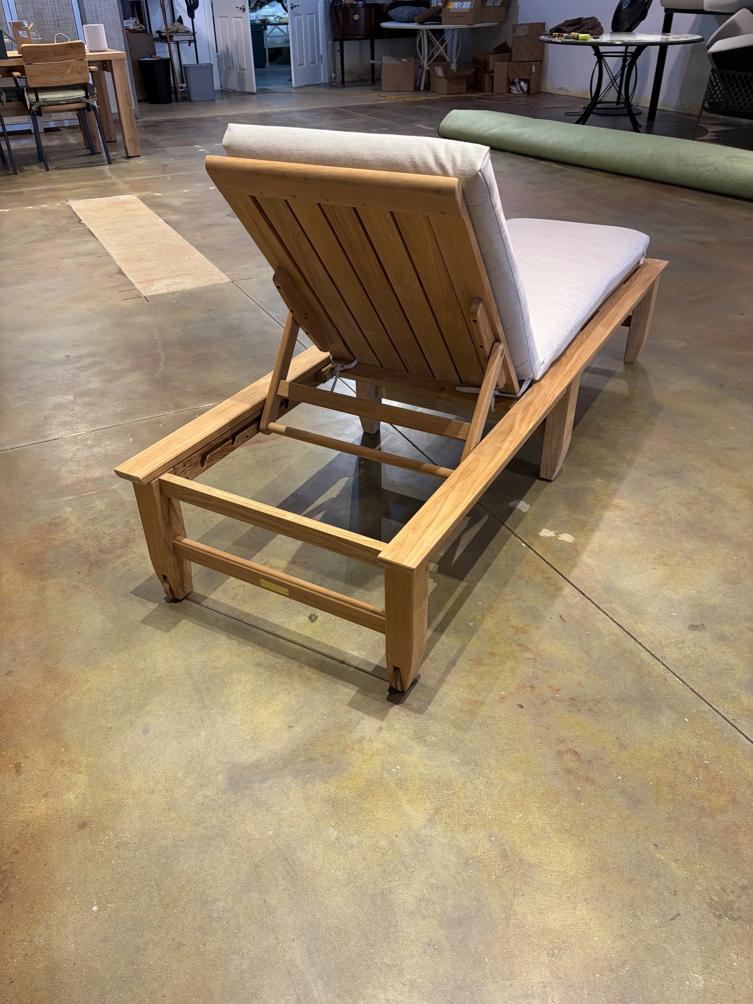 A wooden lounge chair with a light-colored cushion on a frame made of natural wood with a slatted backrest, set on a stained concrete floor inside a room that appears to be a workshop or storage area.