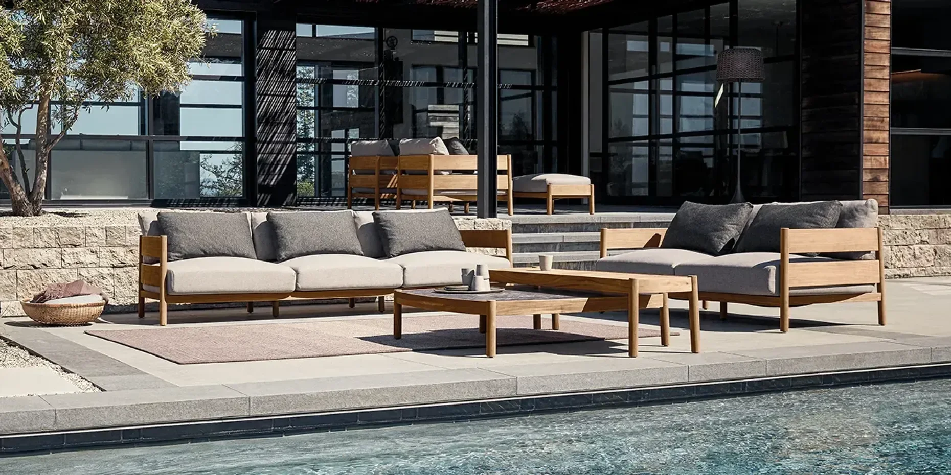 Outdoor patio area with two wooden sofas with gray cushions, a wooden coffee table with cups, a beige rug, steps leading to a modern house with large glass windows, an outdoor lamp, and a potted plant on the left side.