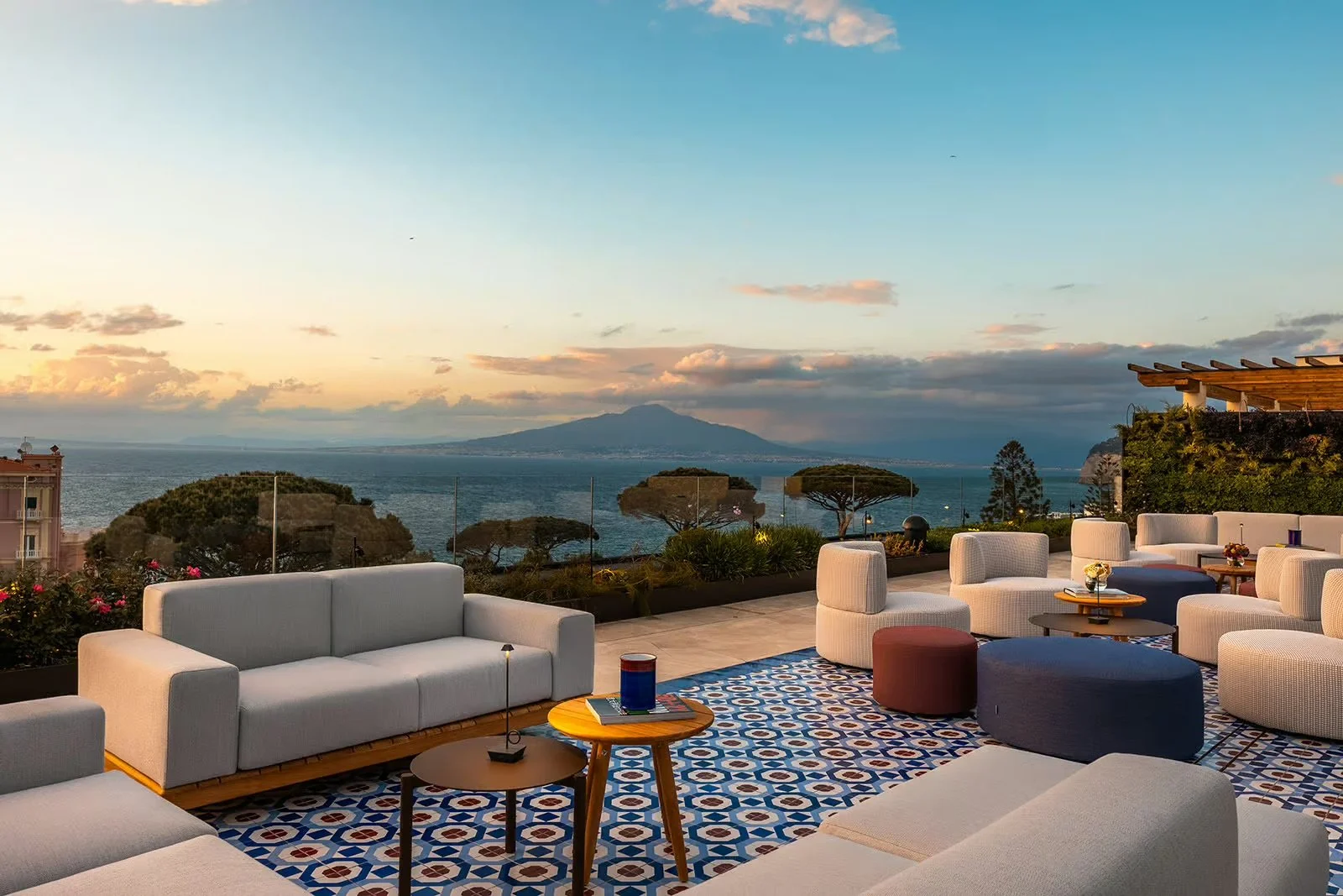 Outdoor seating area with white couches and round tables overlooking the ocean, with a view of a mountain in the distance and sunset sky.