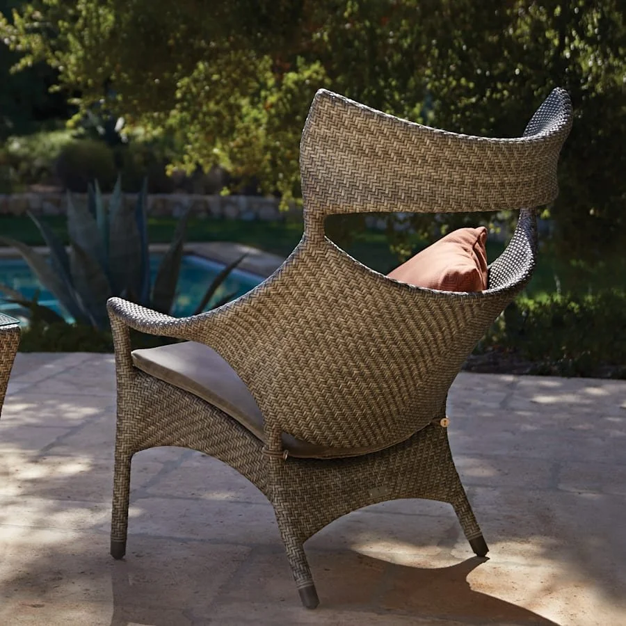Outdoor patio wicker chair with a beige cushion and a brown pillow, overlooking a garden with trees and a swimming pool in the background.