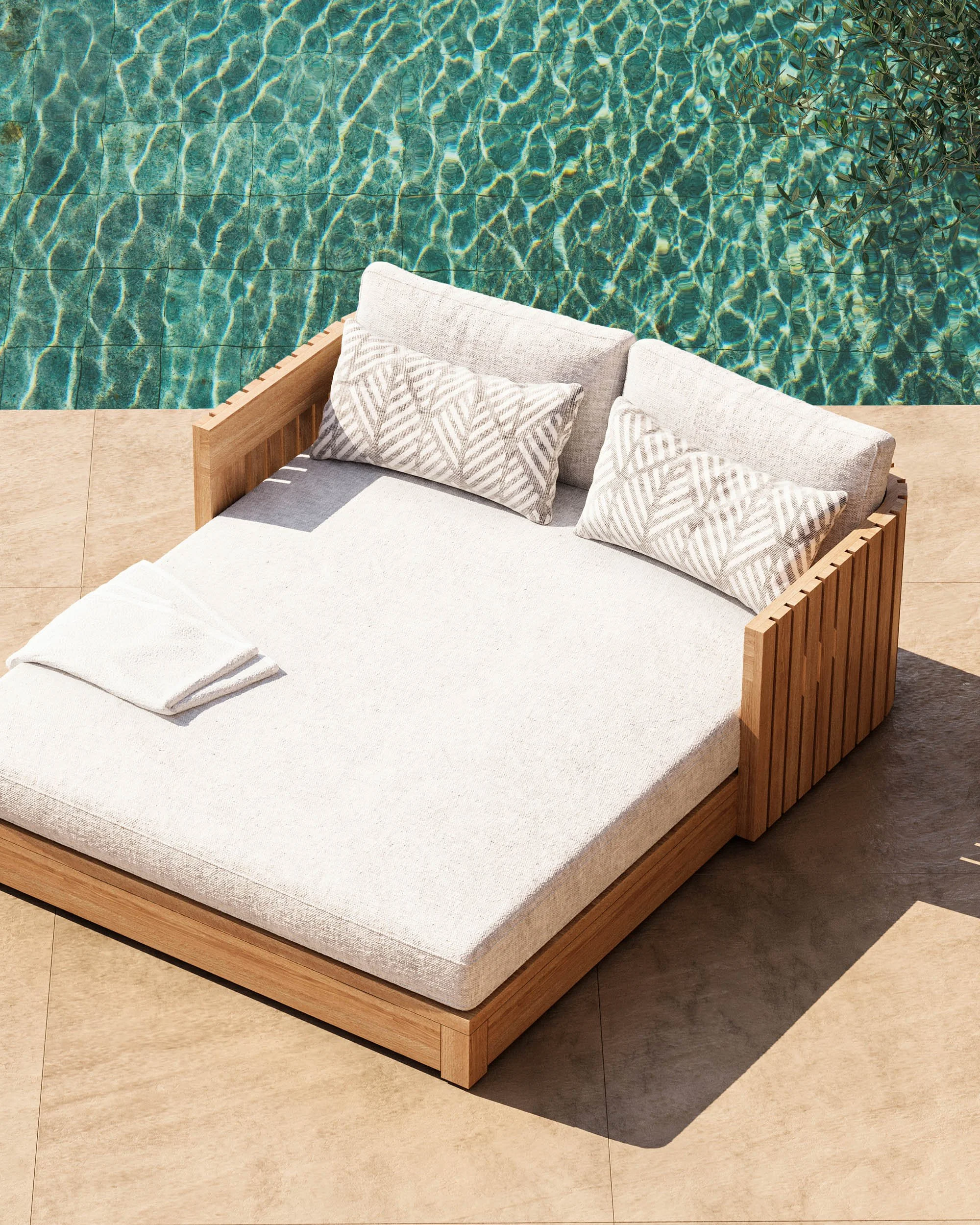 A view of a swimming pool with clear water and sunlight reflections, adjacent to a beige tiled deck with a wooden outdoor bed that has a cream cushion and matching pillows, and a folded towel on top.