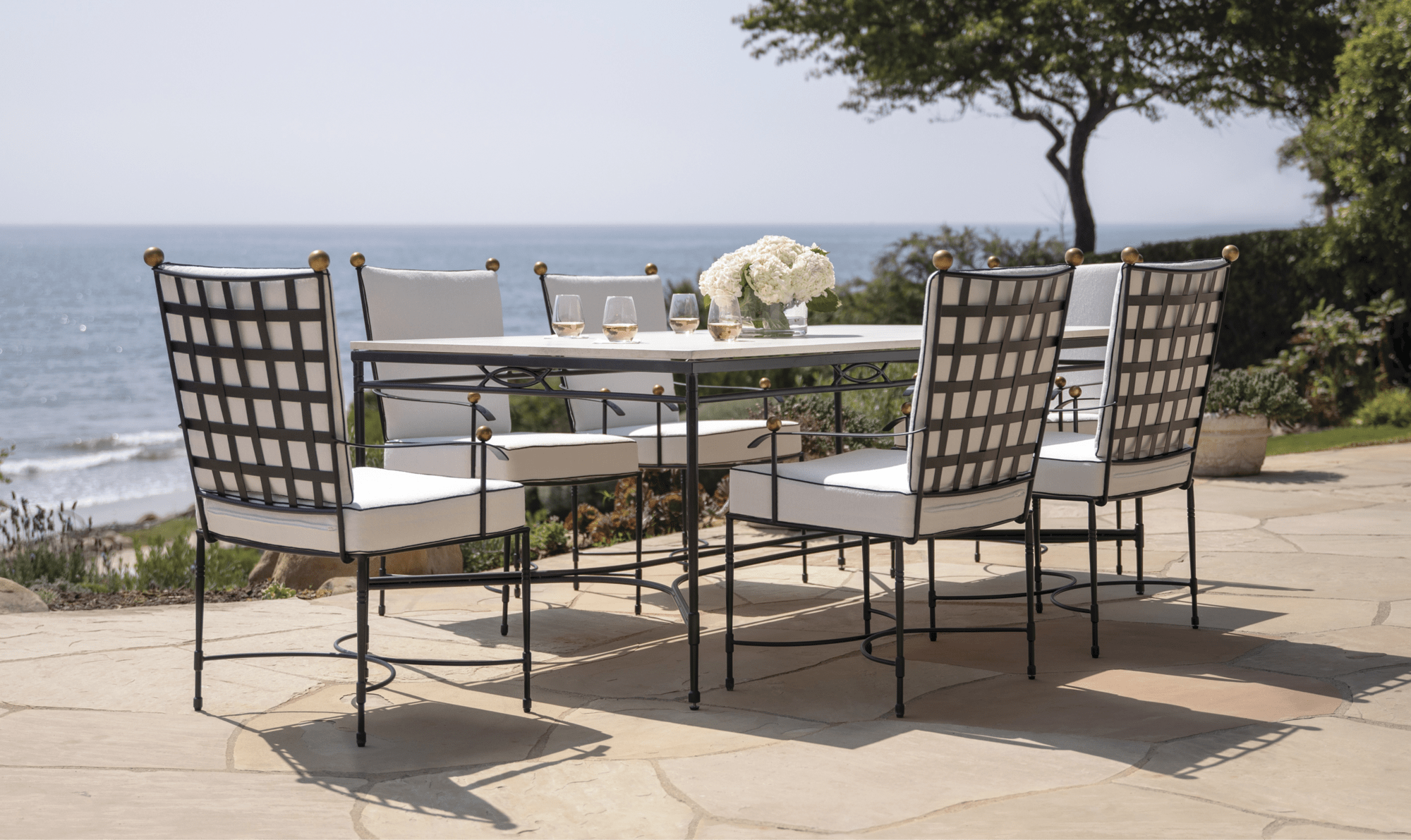 An outdoor patio table with white chairs and a floral centerpiece, overlooking the ocean with trees in the background.