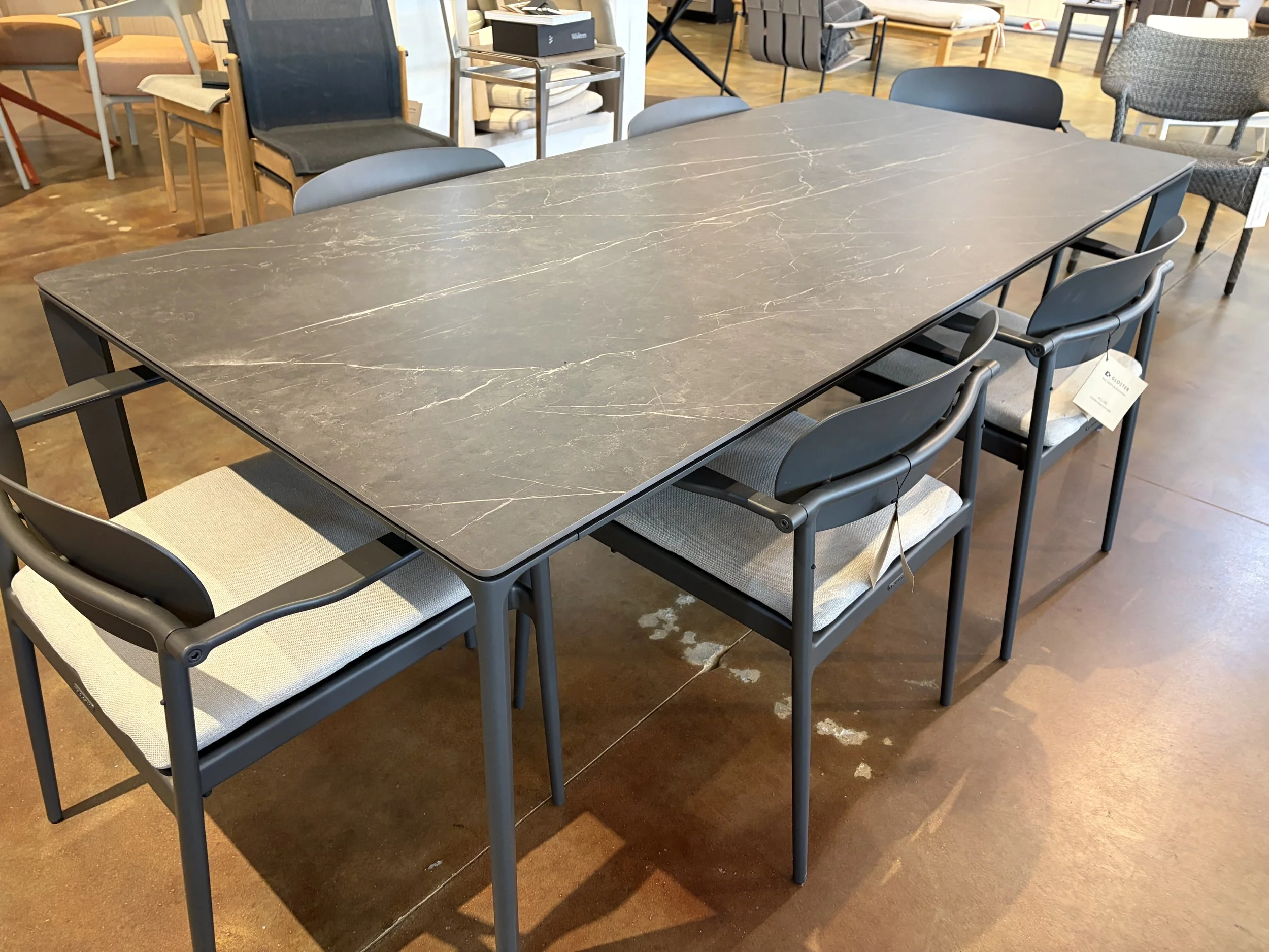 A large rectangular dining table with a dark gray marble-like surface and black edges, surrounded by eight chairs with black frames and light-colored fabric seats, in a furniture store with various chairs and tables displayed around.