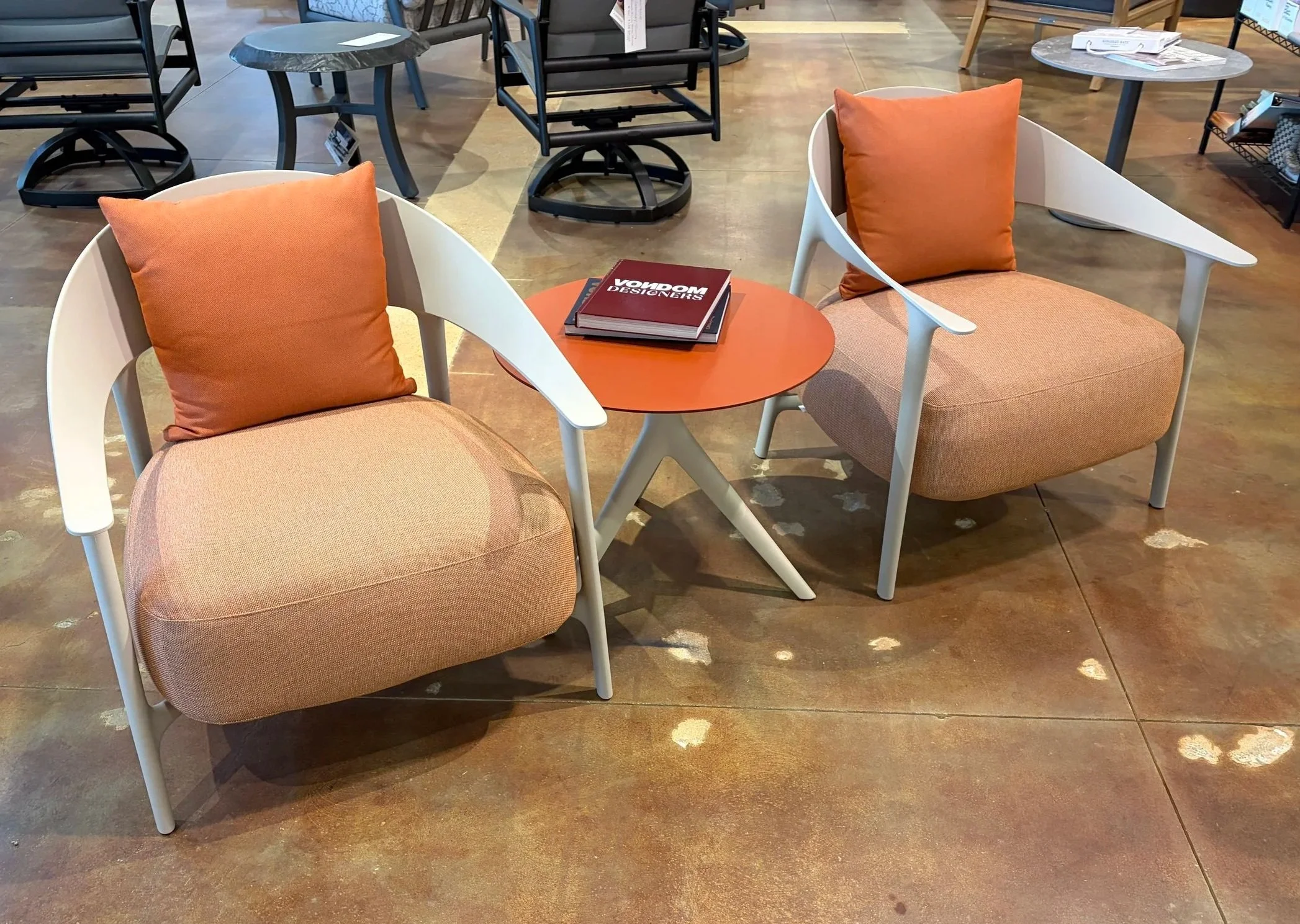 Display of modern furniture in a store, with two chairs featuring orange cushions and armrests, a small round orange table with a magazine on top, and various tables and chairs in the background.