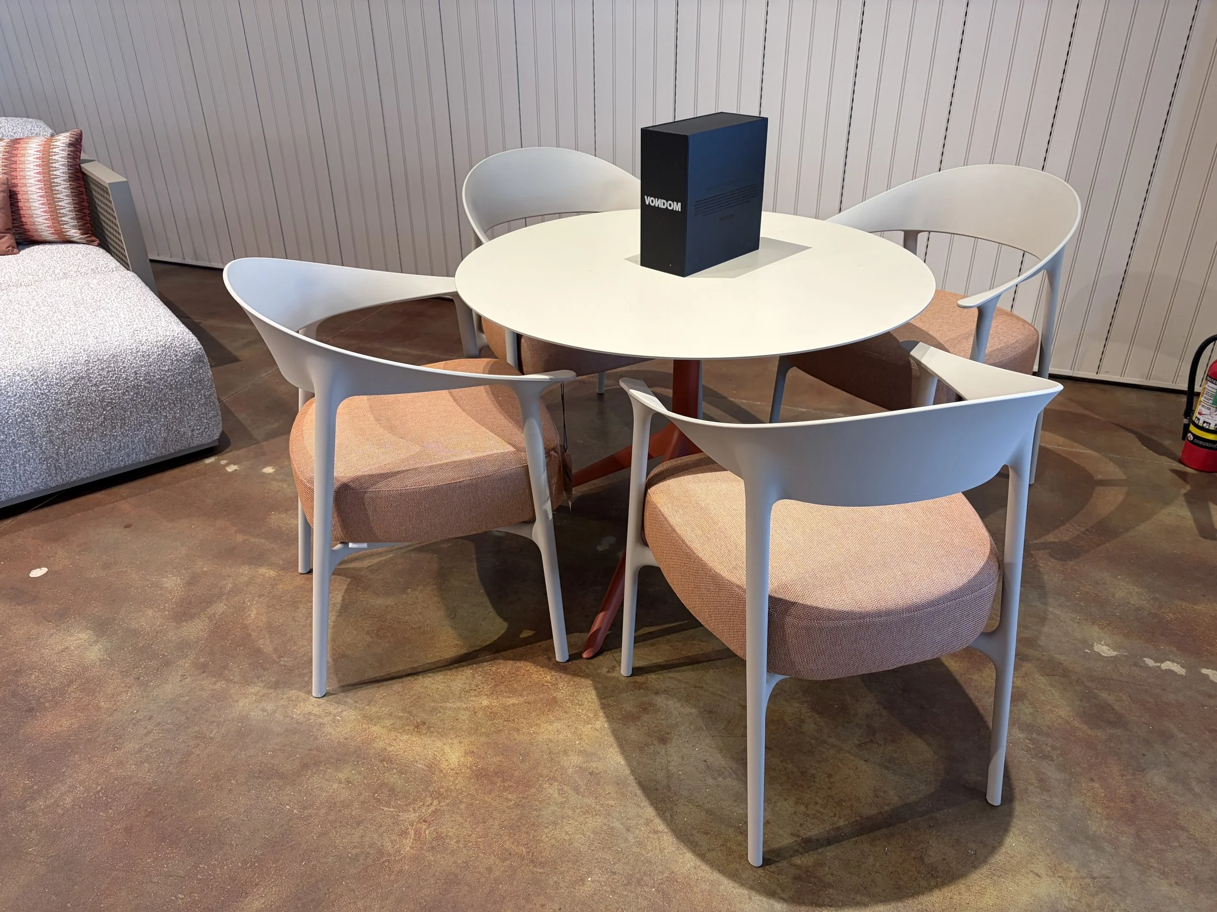 A round dining table with five white chairs, a black box labeled 'MONDO' on top, and a beige sofa with pink and patterned pillows in a room with wooden flooring and a vertical panel wall.
