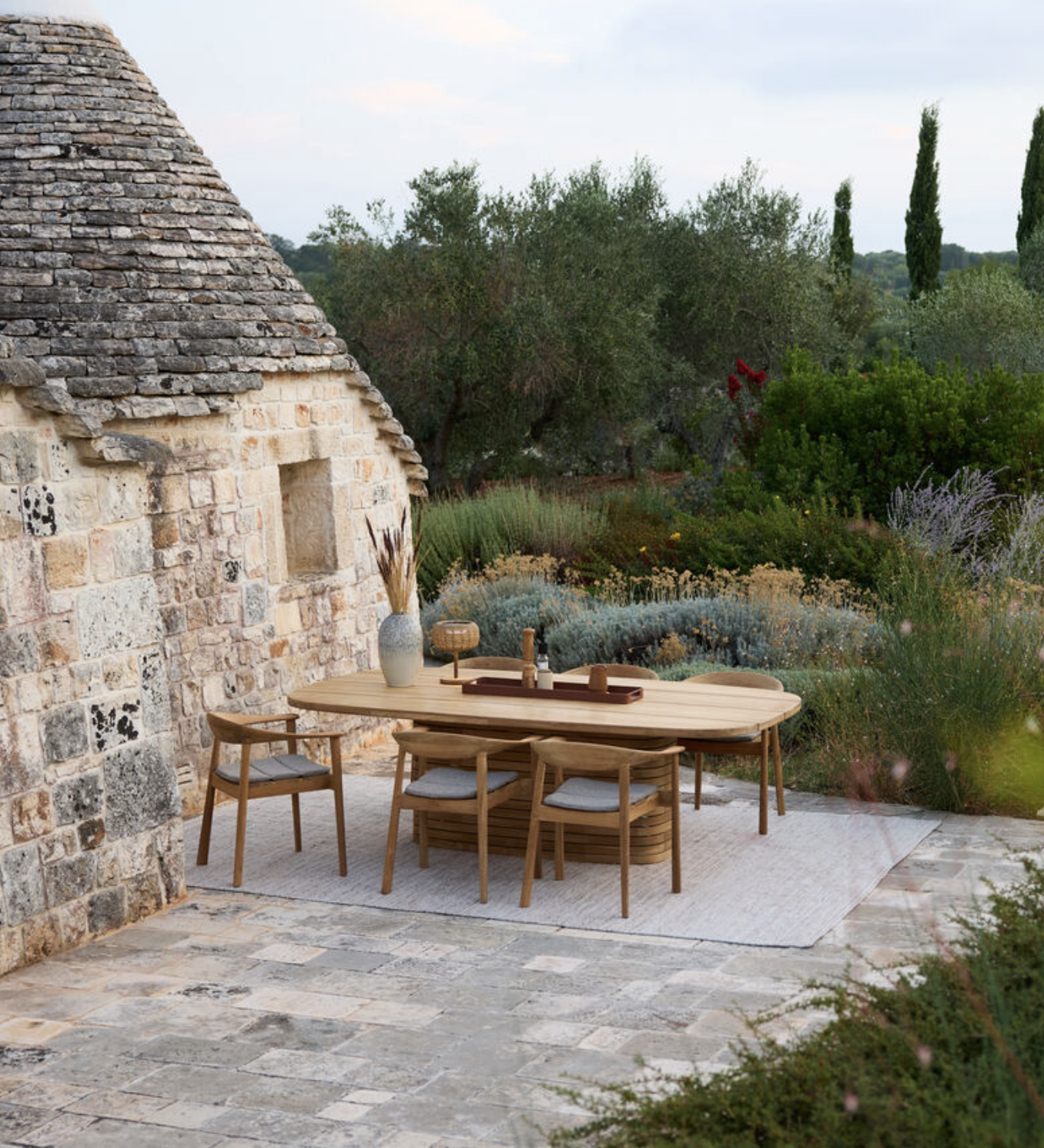 Outdoor dining area with a wooden table, four chairs, a vase, and a tray, surrounded by lush greenery and a stone building wall.