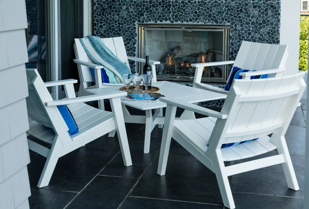 Outdoor patio with a white table and four white chairs, one draped with a striped towel. On the table are a bottle of wine, three wine glasses, and magazines. In the background, there is a modern fireplace with a black pebble surround and a fire inside.