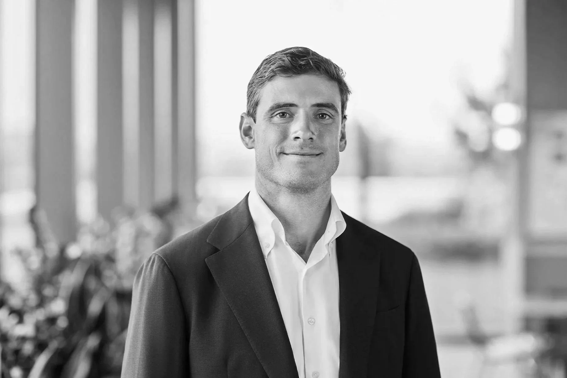Black and white portrait of a man in a suit, smiling slightly, standing in a modern indoor setting with large windows in the background.