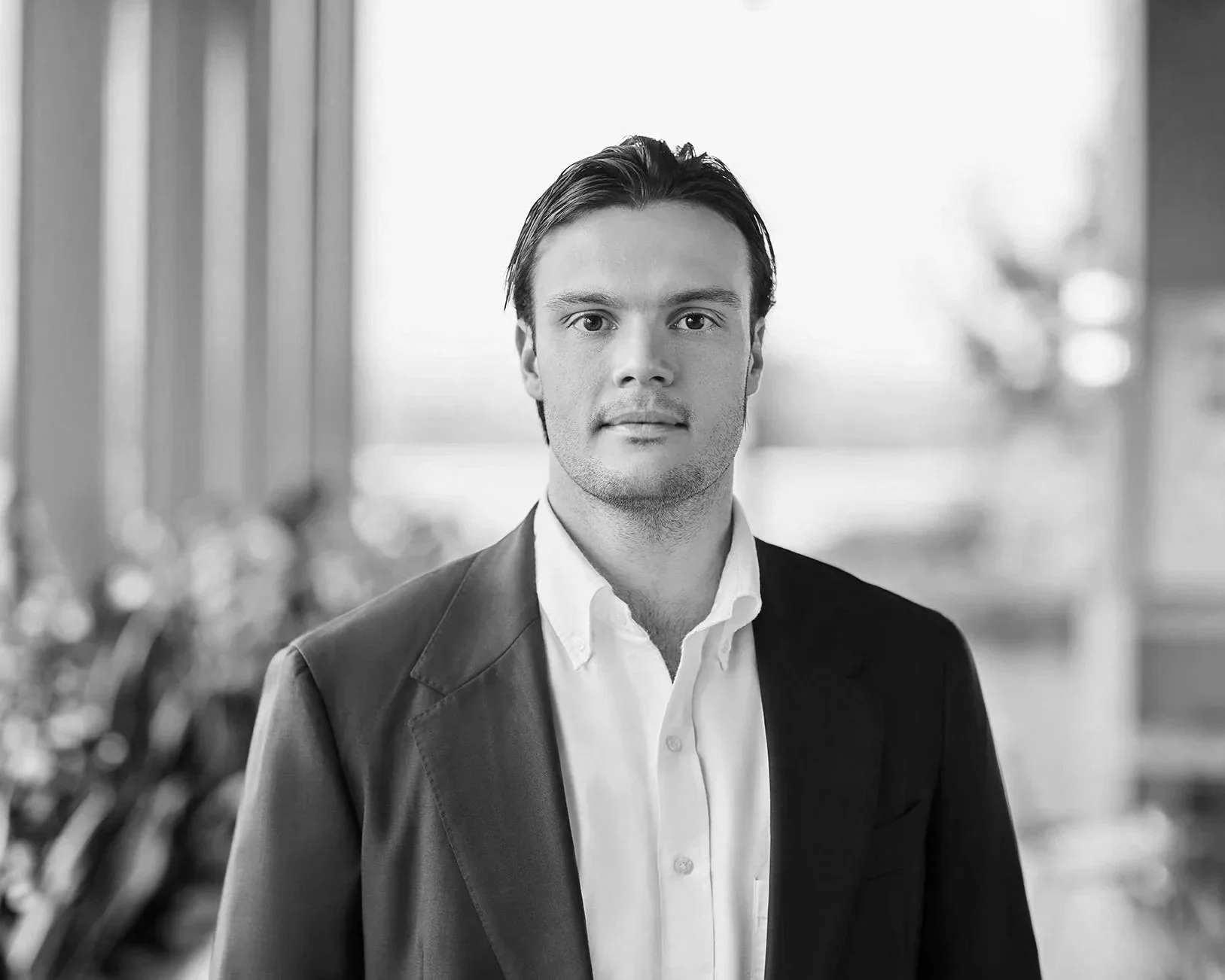 A young man in a suit and white dress shirt standing indoors with a blurred background.