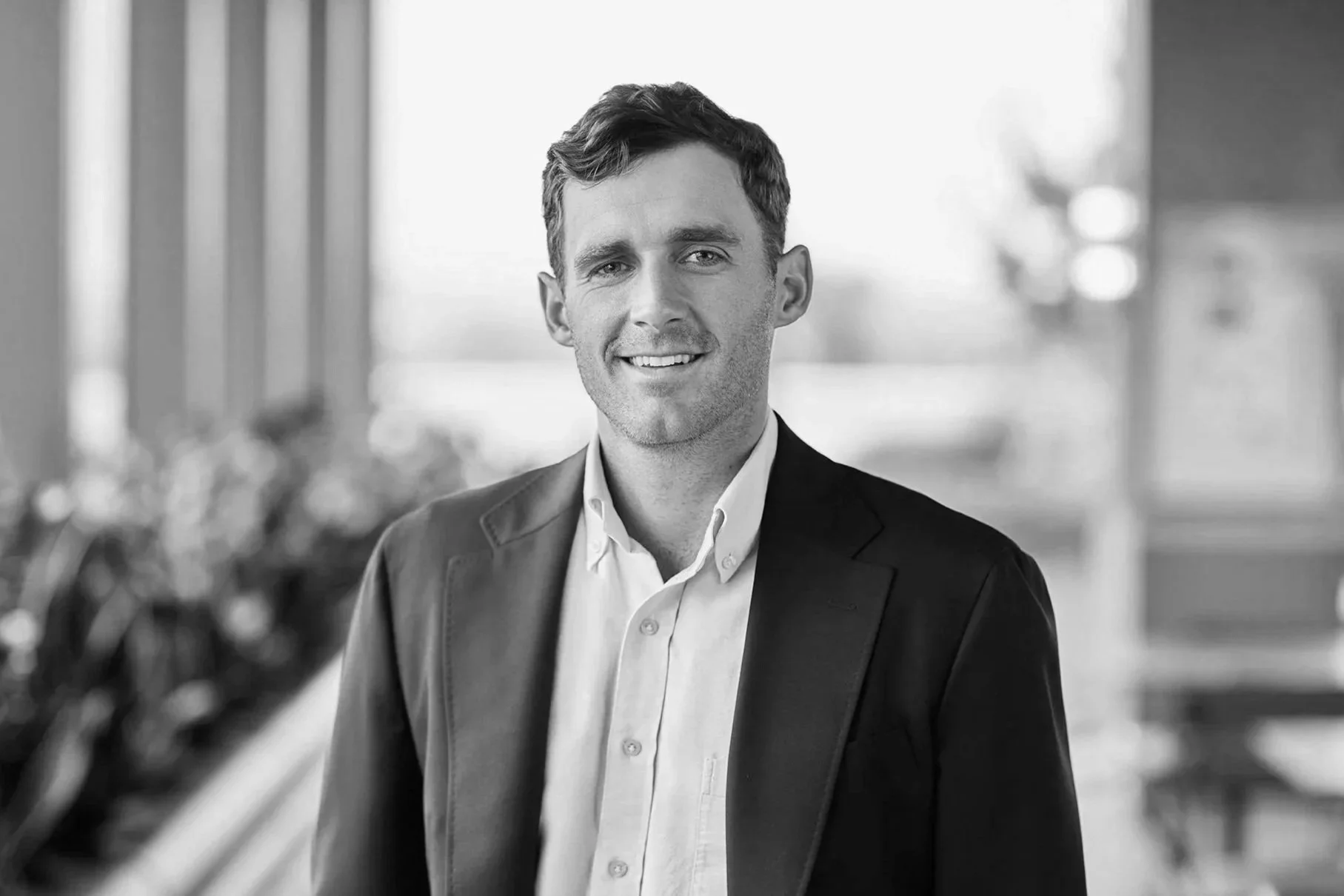 A black and white photo of a smiling man in a suit jacket and white shirt, standing indoors with a blurred background of windows and outdoor scenery.