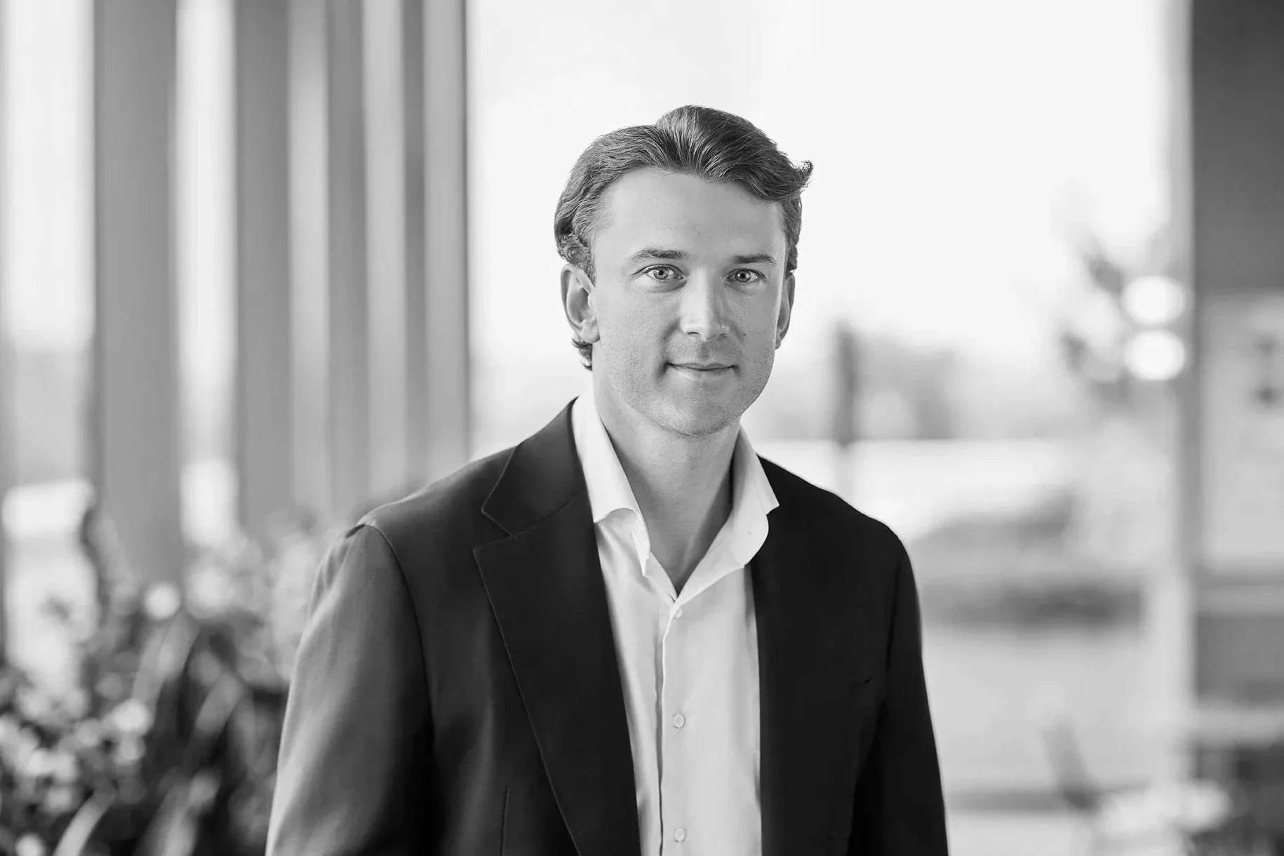 Black and white photo of a man in a suit and white shirt standing indoors near large windows with a blurred cityscape in the background.