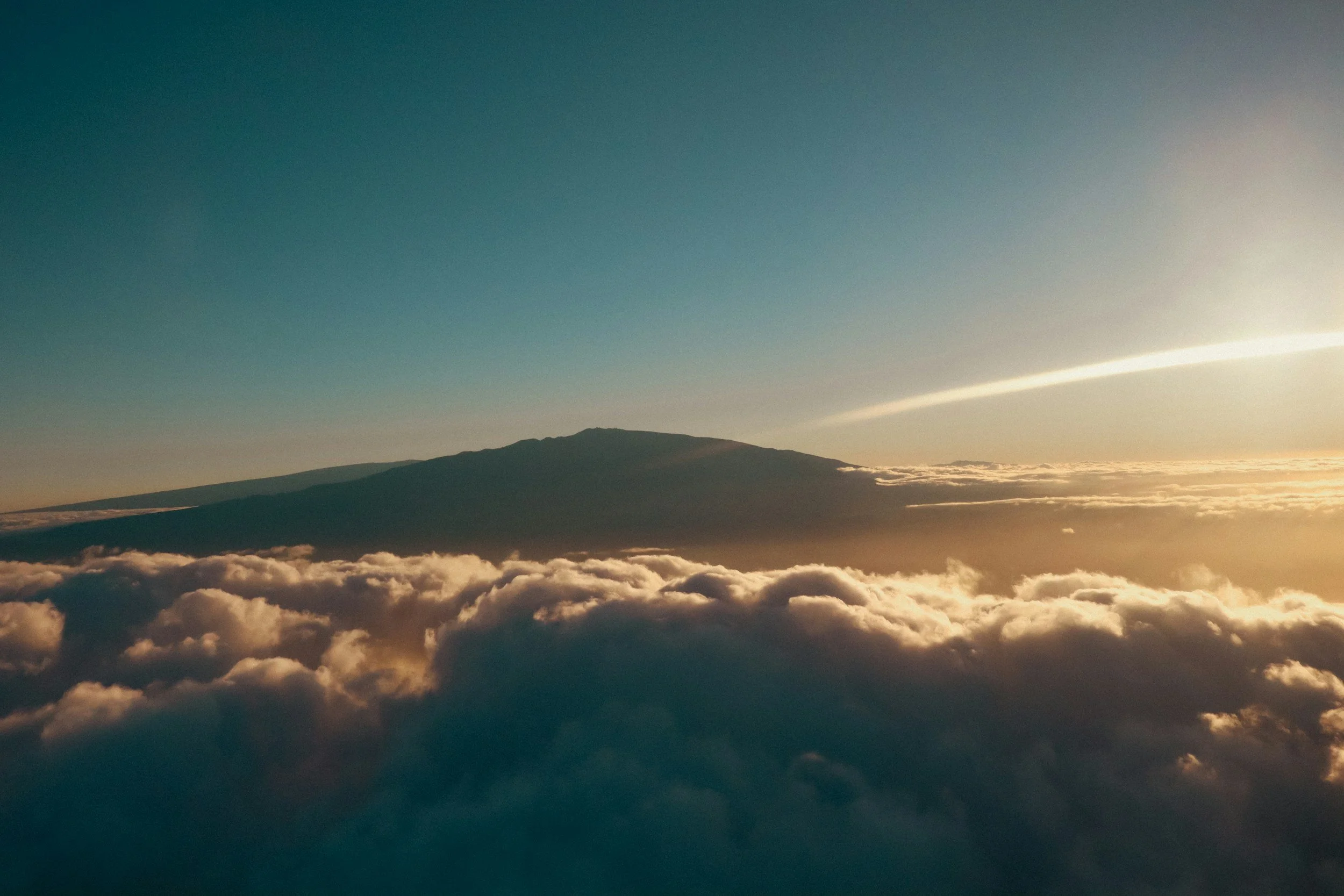 An aerial view of a mountain peak above a dense layer of clouds during sunset or sunrise.