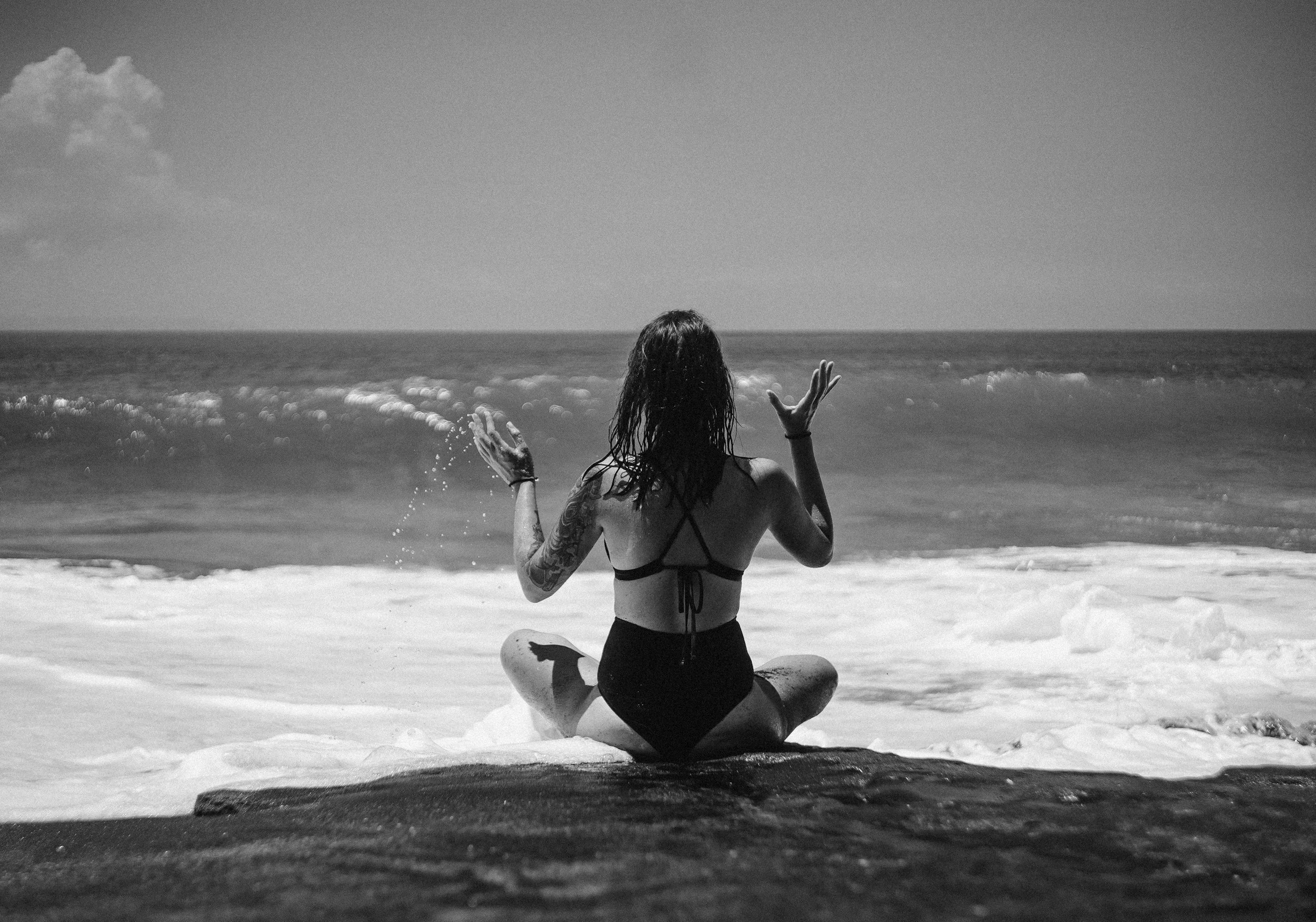 A woman sitting on a rock at the beach, facing the ocean, with her hair wet and wearing a bikini, raising her hands during daytime.