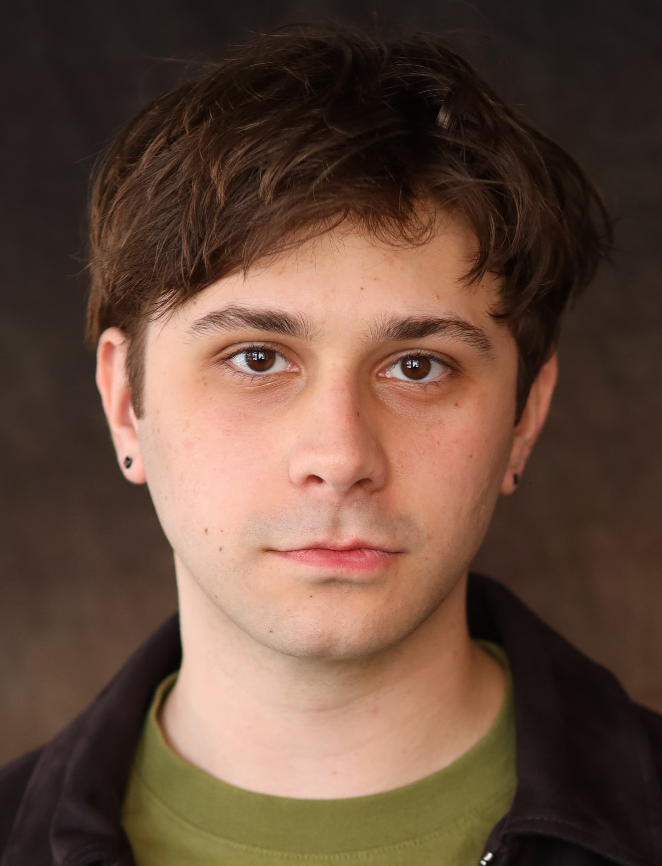 Close-up of a young person with short, dark brown hair, brown eyes, and light skin, wearing a green shirt and a black jacket.