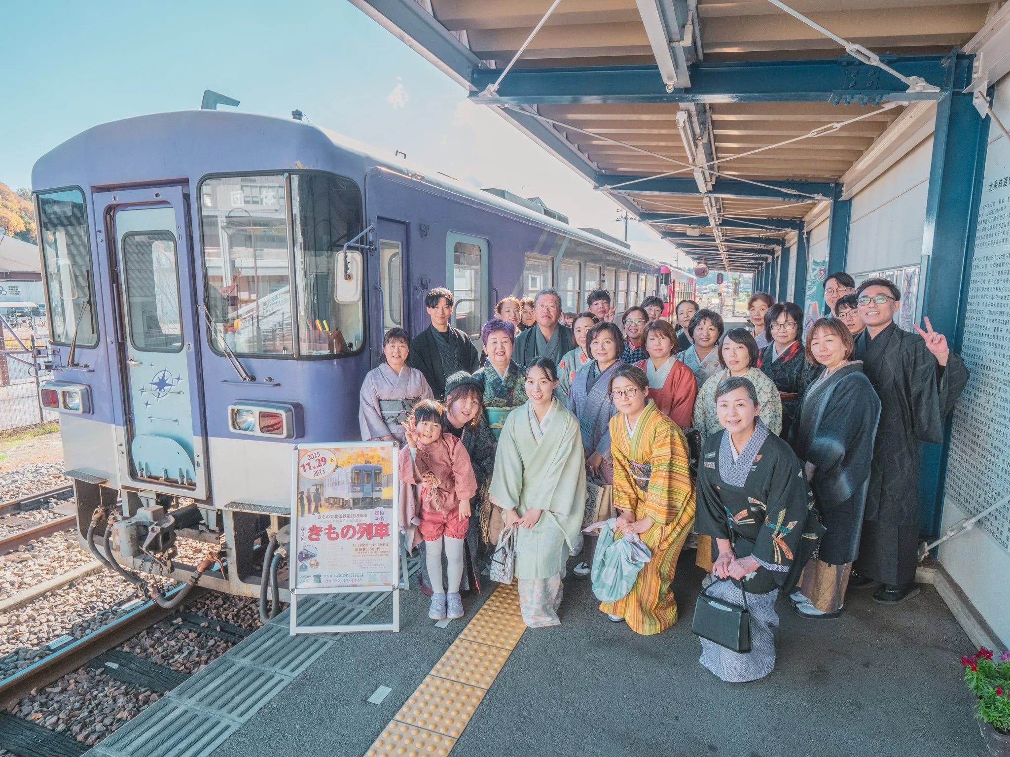 多くの人々が伝統的な着物を着て、青い列車の前で写真を撮っている。列車と駅のプラットフォームが見える。