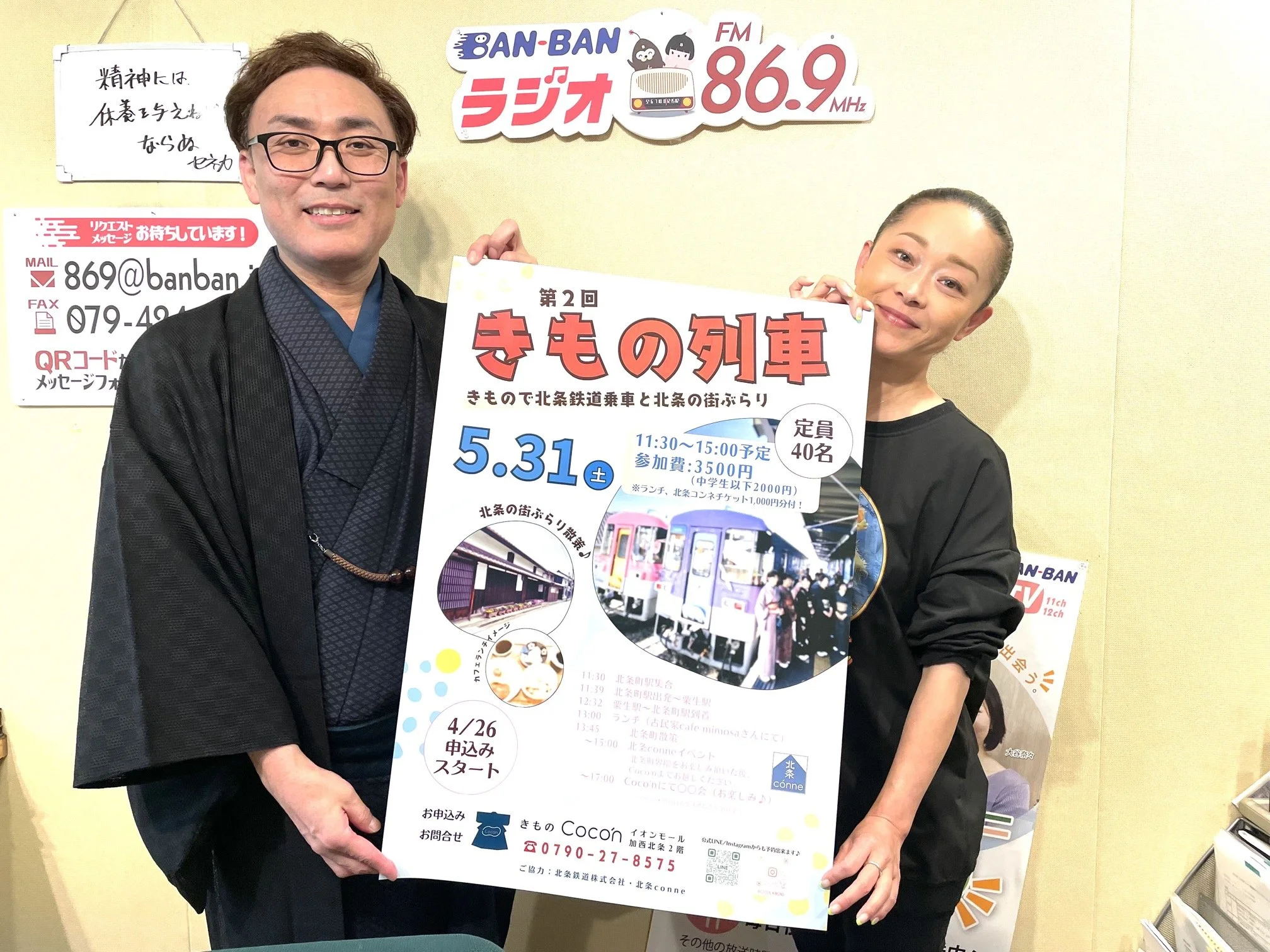 二人の人物がイエローカラーのポスターを持って立っている。背景にはラジオ局の看板とその他の掲示物が見える。