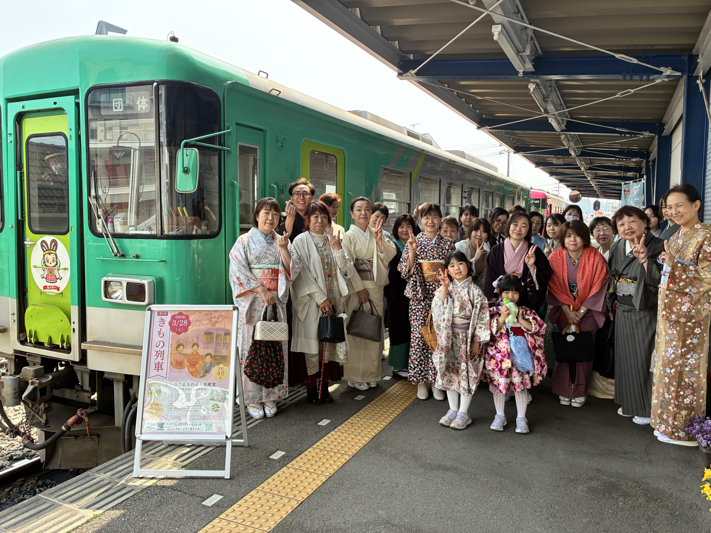 着物を着た家族や友人たちが、駅のホームで記念撮影をしている。背景には緑色の列車が停車している。皆笑顔でポーズをとっている。