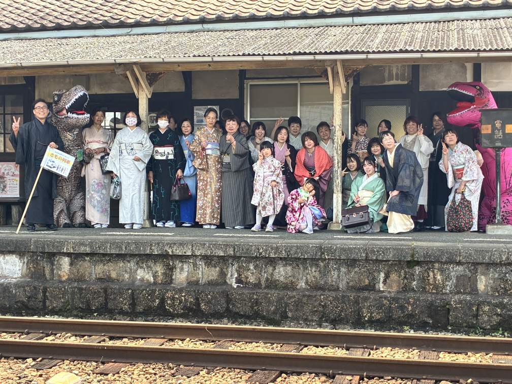 伝統的な着物や浴衣を着た人々が駅のプラットフォームでグループ写真を撮っている。有人の駅舎、ダイナミックな恐竜の着ぐるみも見える。