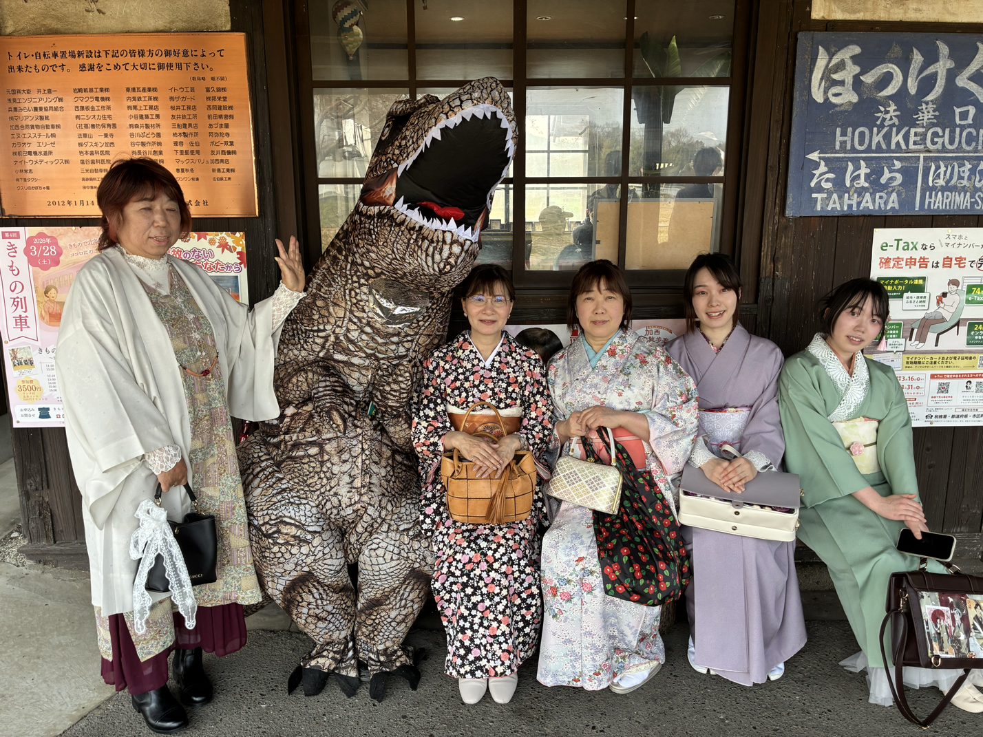 着物を着た女性五人と中間に恐竜の着ぐるみを着た男性が並んでいる写真。背景には看板や窓が見える。