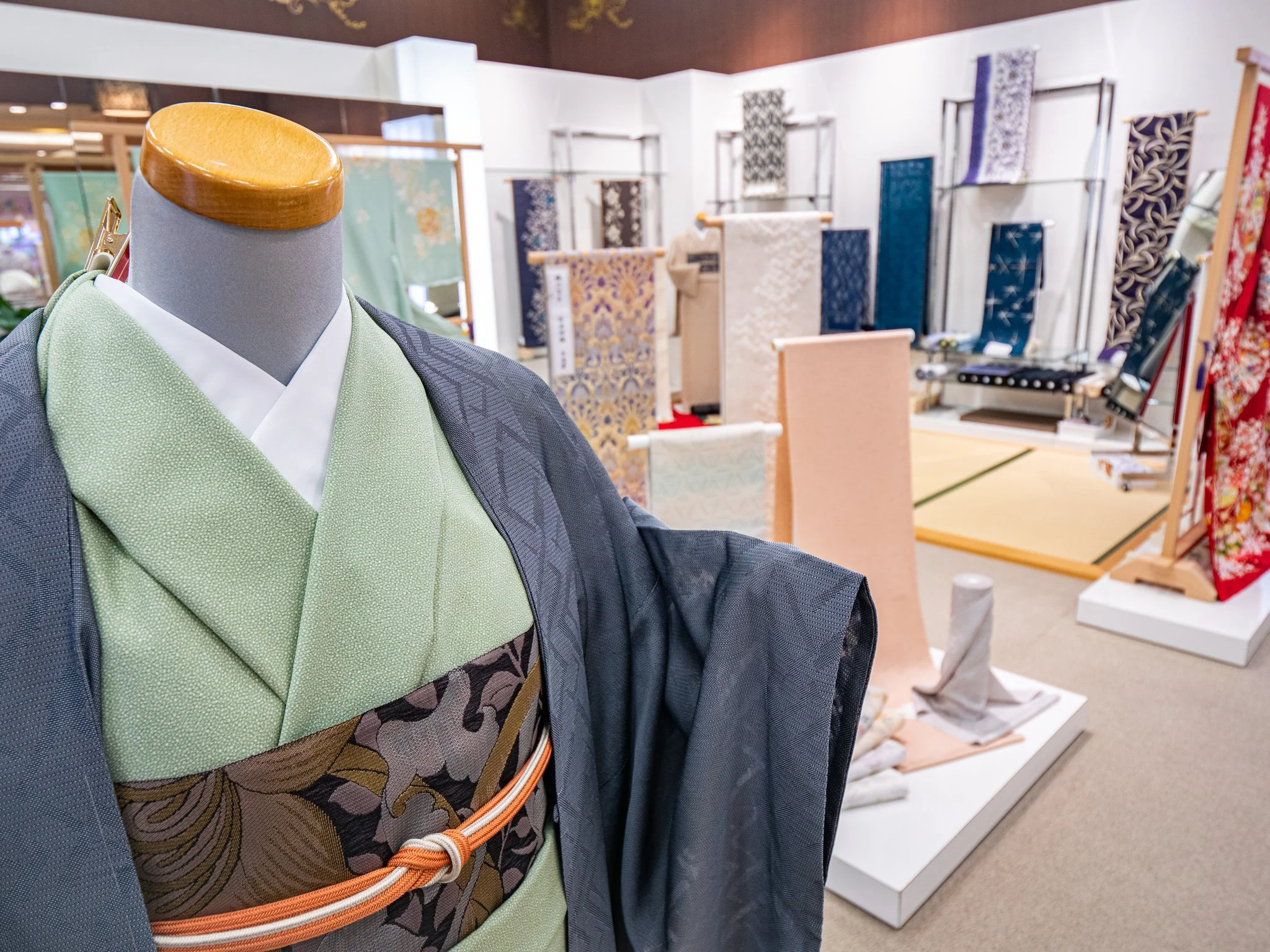 着物の展示販売コーナーで、前景に緑色の着物を着たマネキンが映っている。背景には様々な色と柄の着物や帯が展示されている。