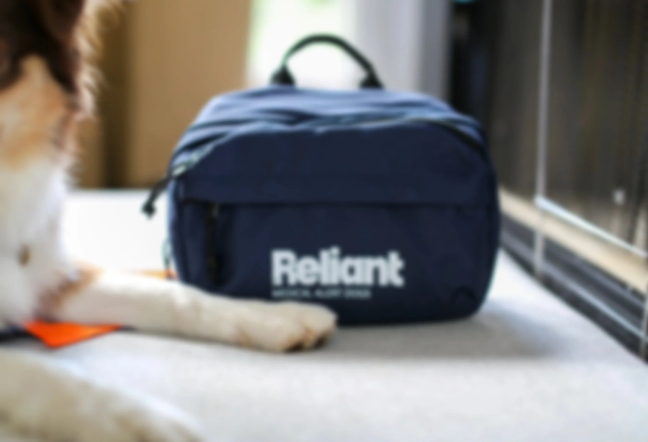 Blurred image of a blue Relief backpack placed on a light-colored surface, with part of a person's hand and arm visible in the foreground, and a small orange object near the bottom left corner.
