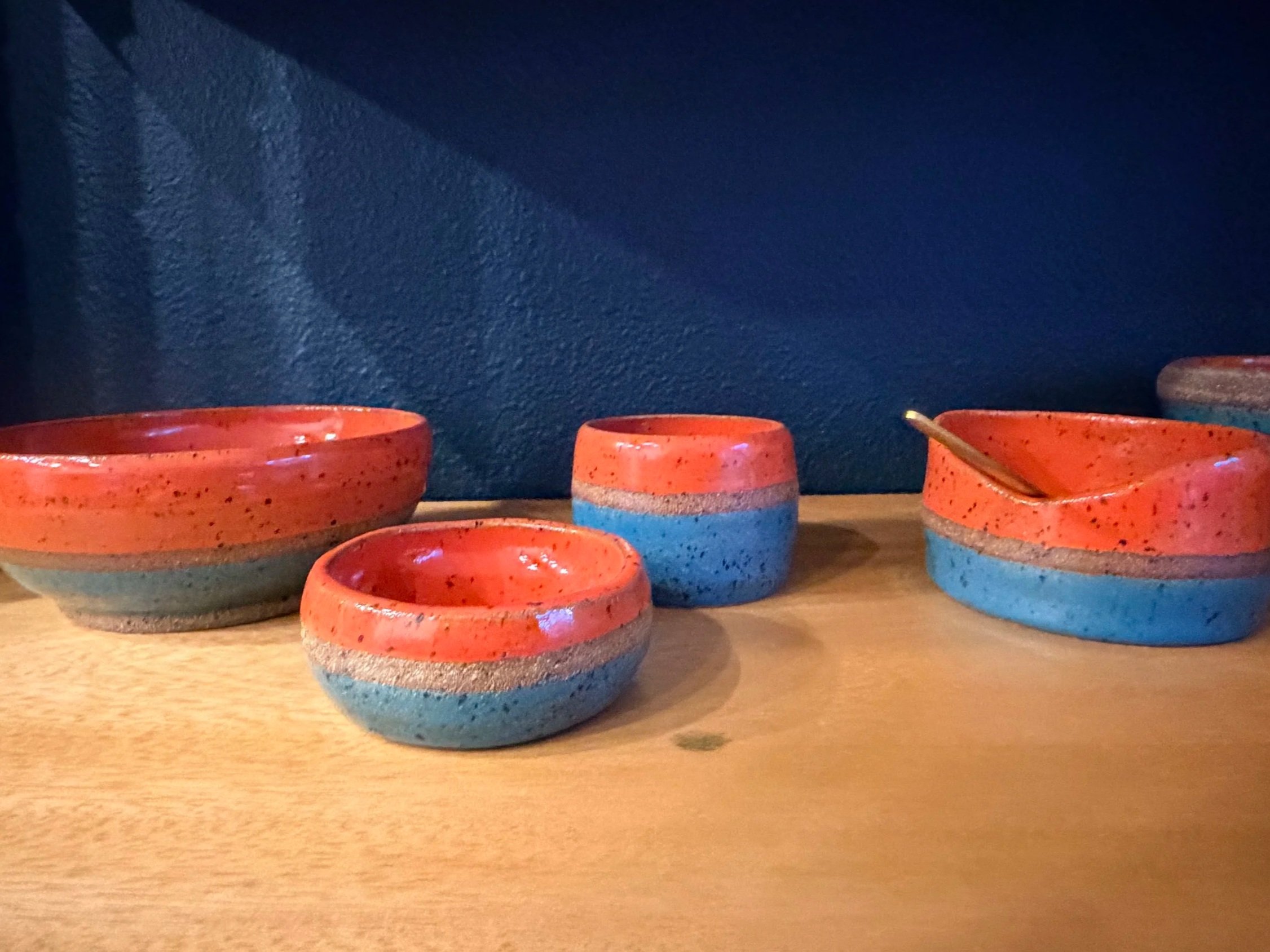 Set of four colorful ceramic bowls and cups with orange, blue, and pink glaze, placed on a wooden surface against a dark blue wall.