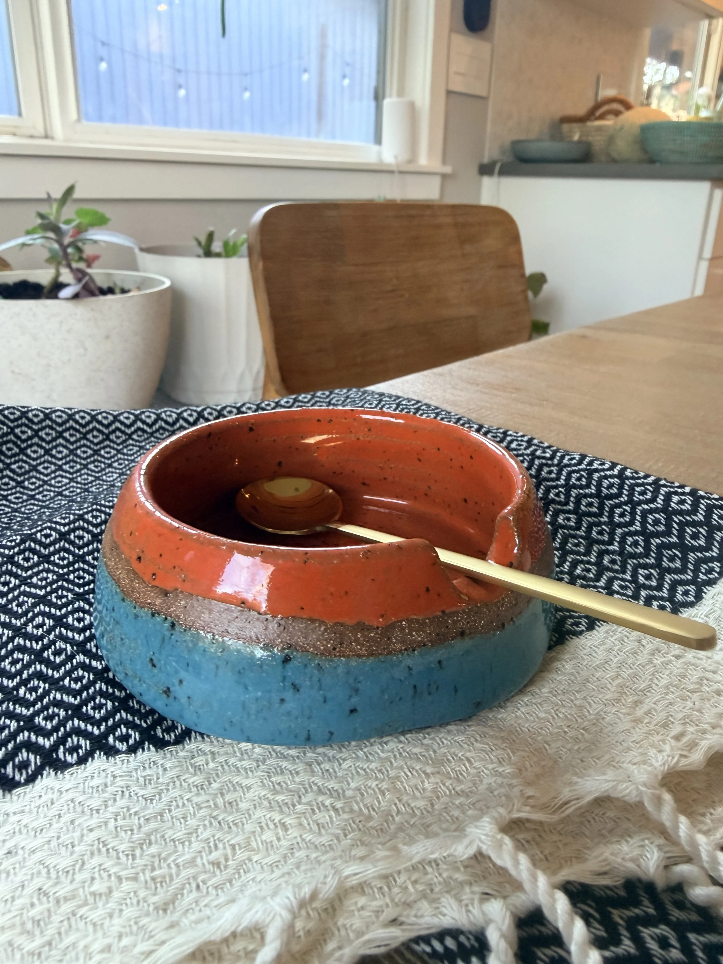 Colorful ceramic salt dish on a table with a golden spoon resting inside, set on a black-and-white patterned cloth with a beige cloth underneath, in a cozy kitchen or dining area with chairs, plants, and window in the background.