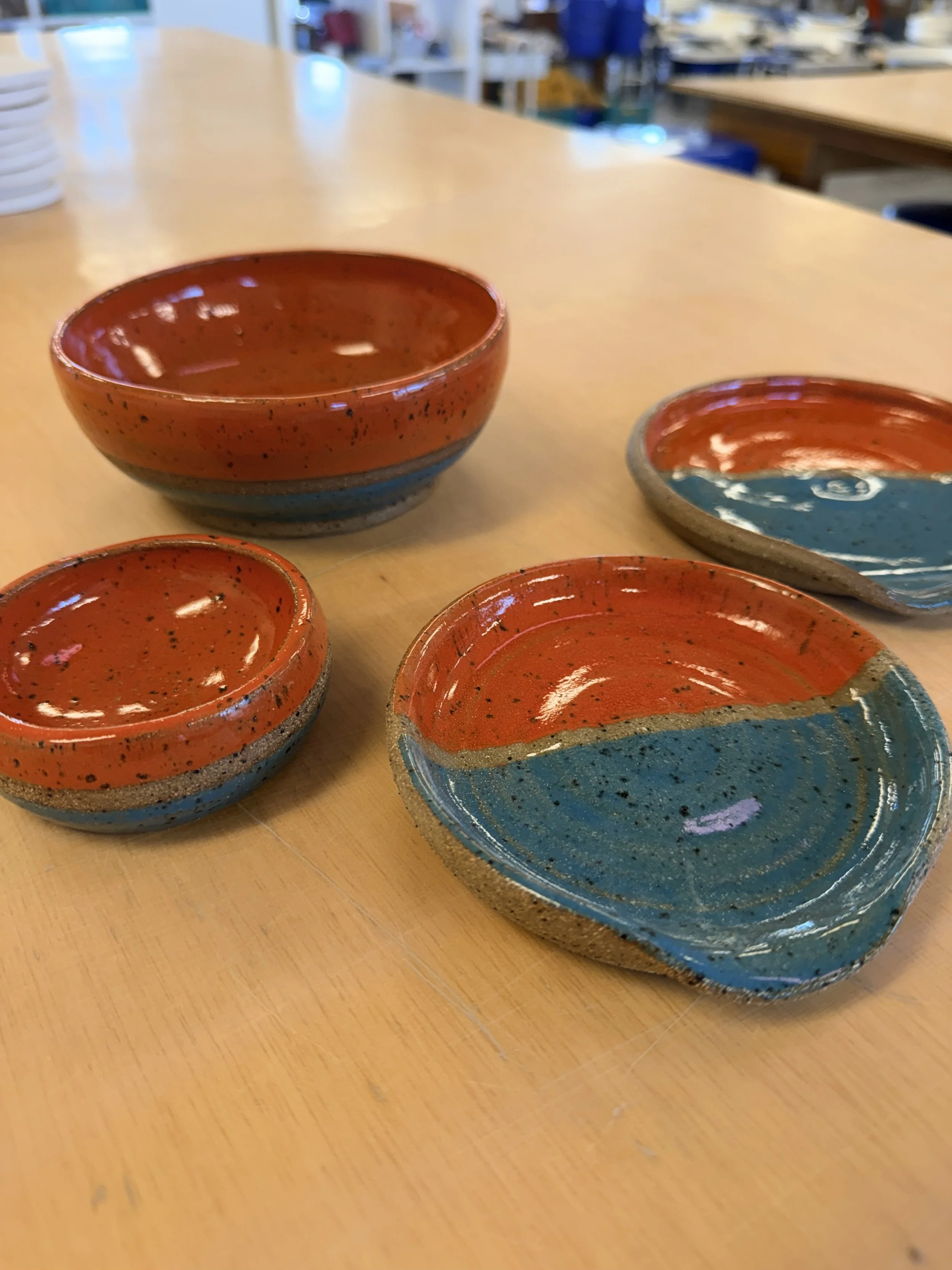 Set of four handmade, glazed ceramic bowls with teal and tomato patterns on a wooden table in a ceramics studio.