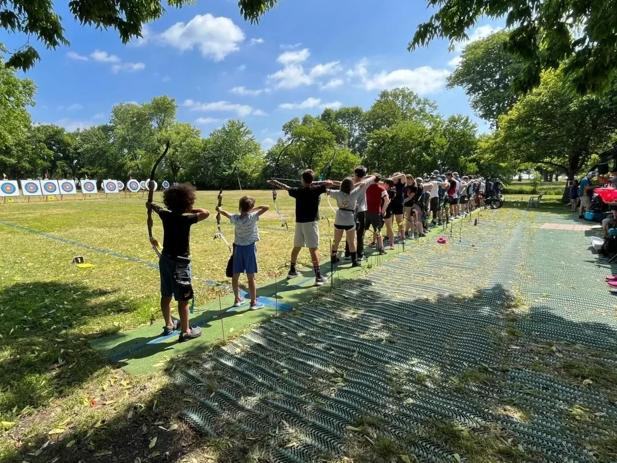 A group of children and adults practicing archery outdoors on a sunny day, lined up and aiming bows at targets in a park.