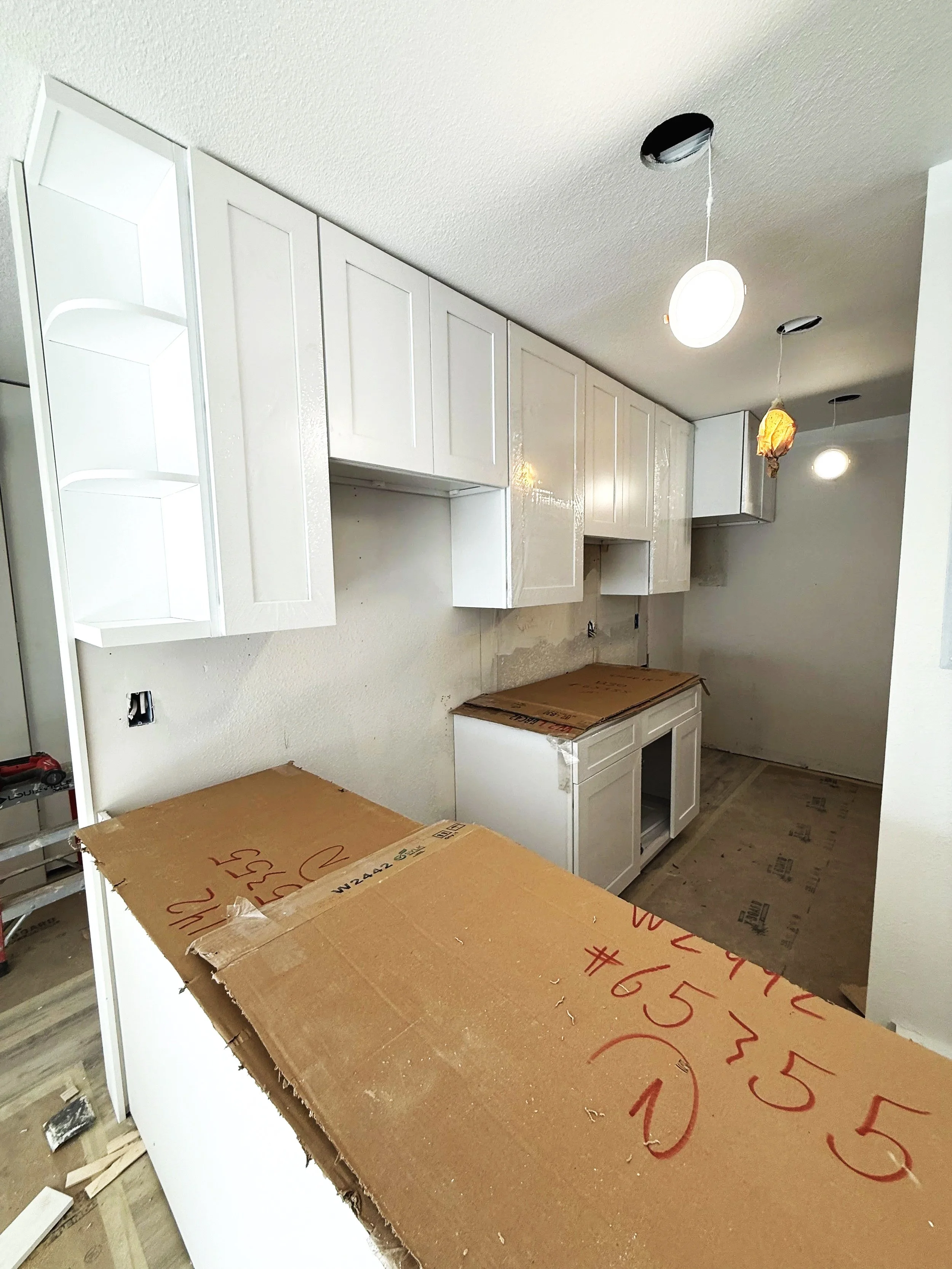 Kitchen under construction with white cabinets, a large cardboard box on a counter, and exposed lighting fixtures on the ceiling.