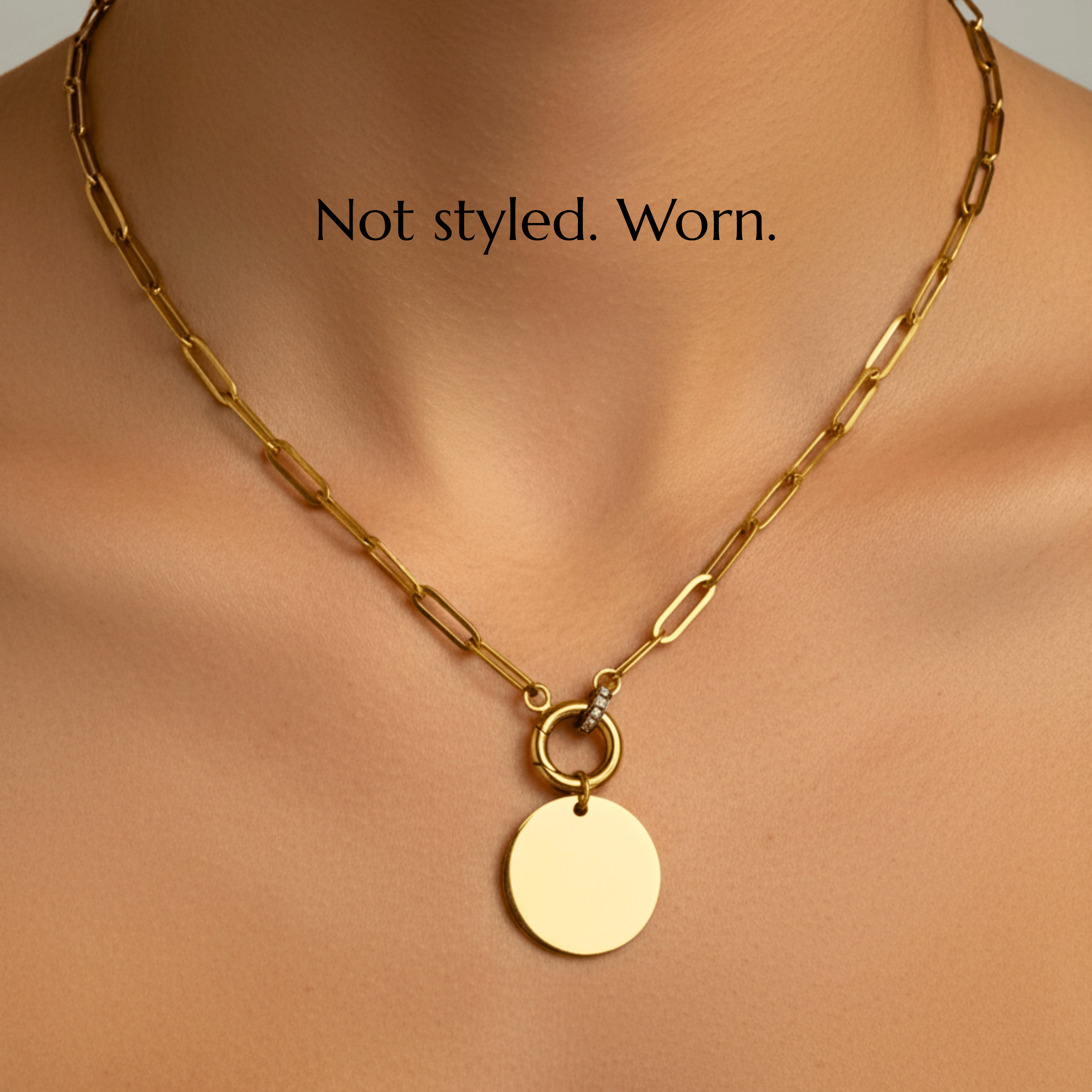 Close-up of a gold paperclip chain necklace with a circular pendant, worn on a person's neck, with the text 'Not styled. Worn.' superimposed.