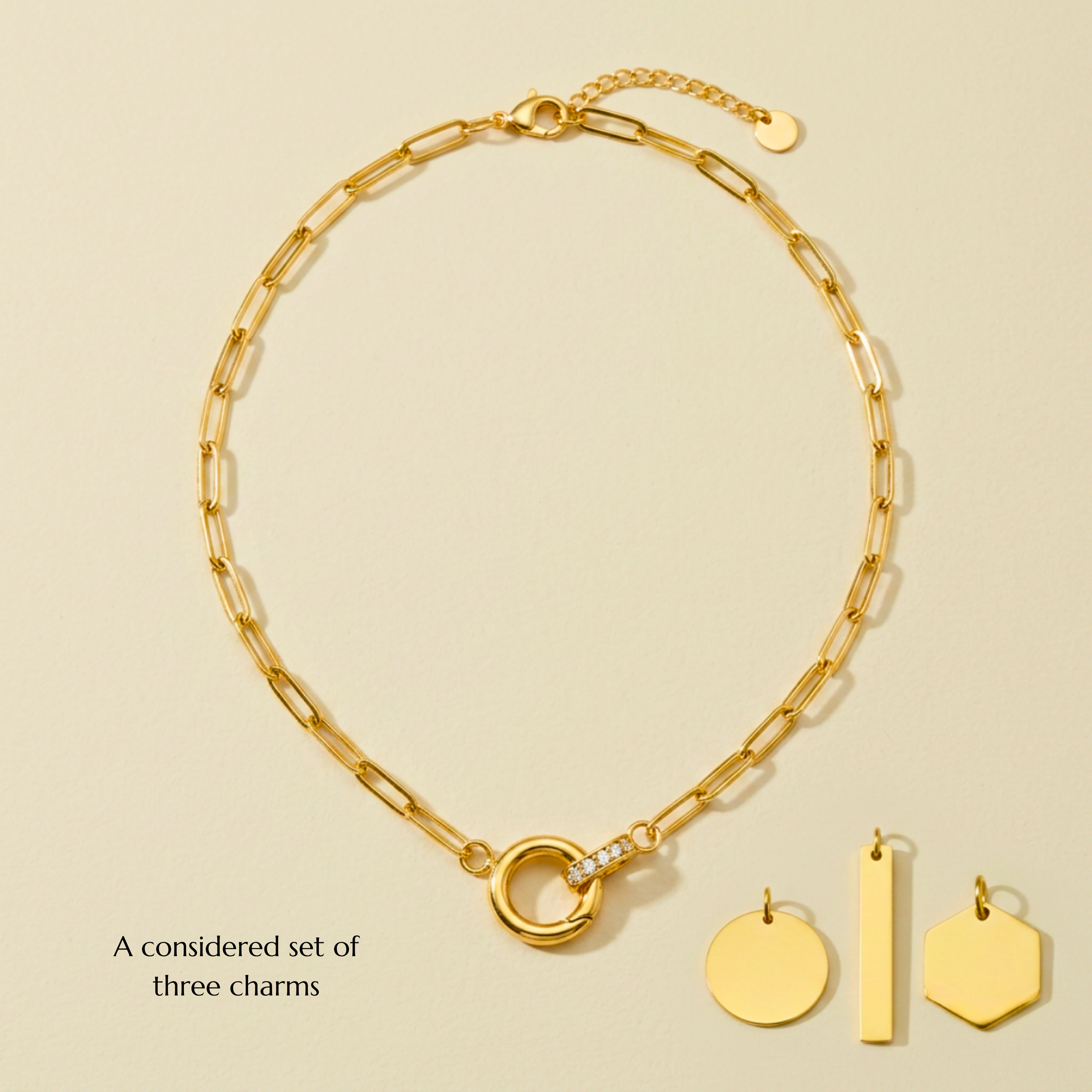A collection of gold jewelry including a chain necklace, a pendant necklace with a semi-circular charm encrusted with small stones, and three gold charms with a circle, rectangle, and irregular shape, hanging on a light-colored background.