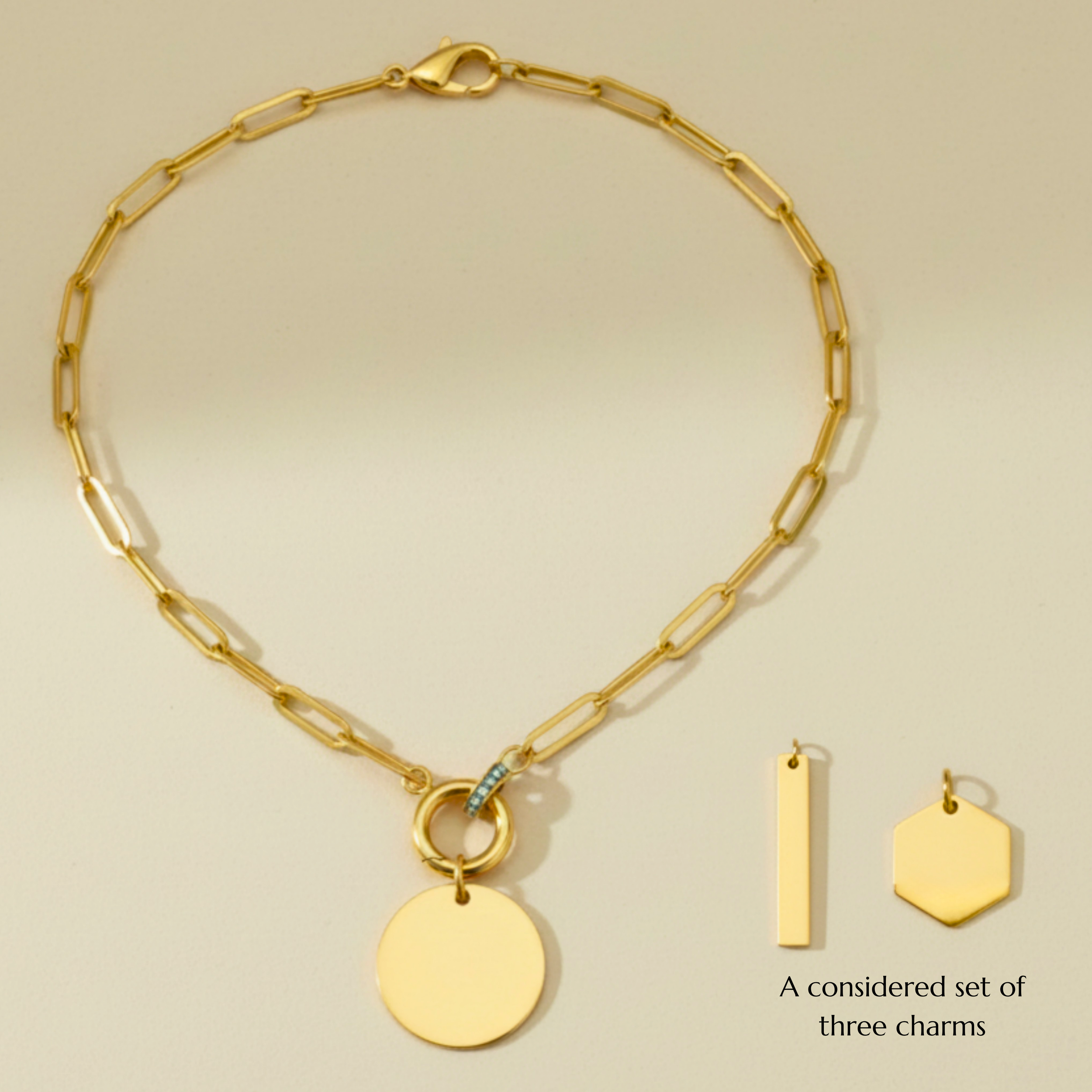 A gold paperclip necklace with a round charm pendant and two gold charms, one rectangular and one hexagonal, laid out on a neutral surface.