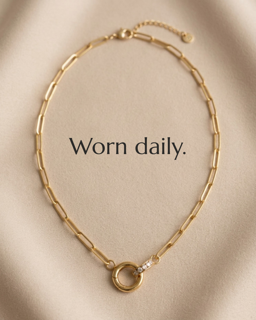 Gold chain necklace with a circular pendant and small diamonds, on a beige fabric background with the words 'Worn daily.'