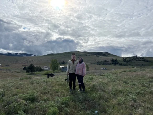 Two people standing close together in an open grassy field with a hilly landscape and cloudy sky in the background.