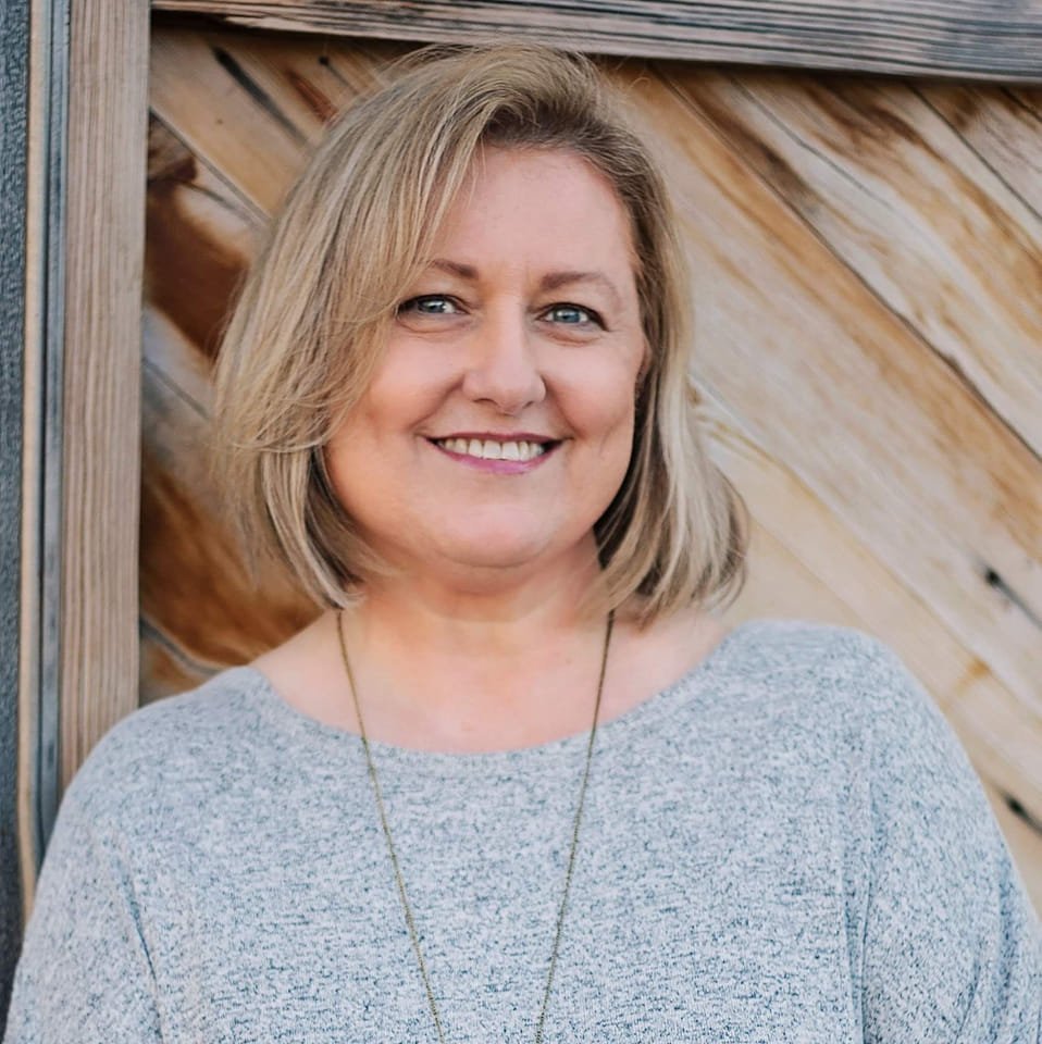 A woman with shoulder-length blonde hair smiling, standing outdoors against a wooden background.