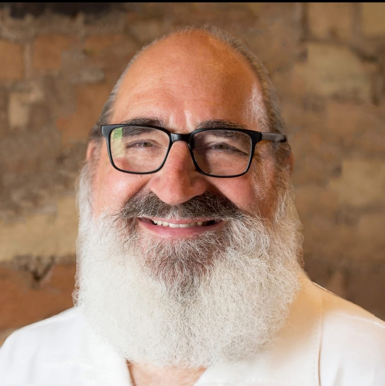 A smiling man with a full white beard, glasses, and a bald head, standing in front of a brick wall.