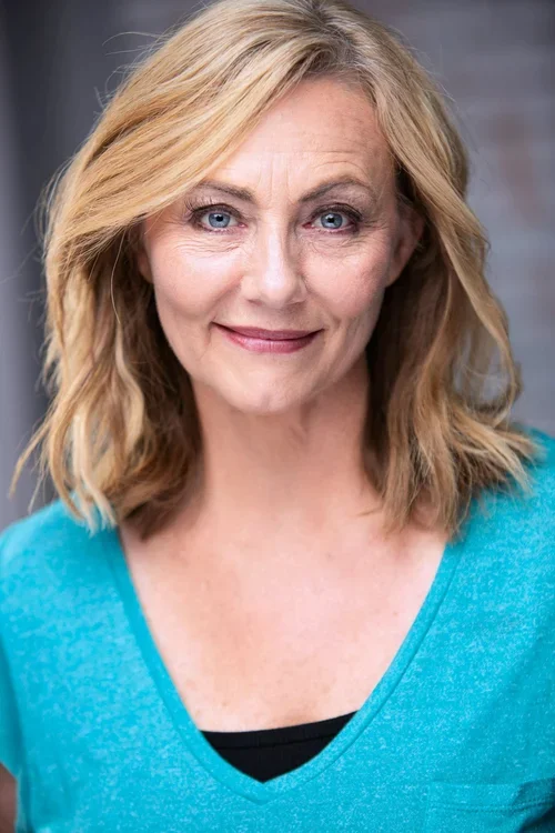 A middle-aged woman with shoulder-length blonde hair, winking, smiling, wearing a turquoise shirt, standing in front of a gray background.