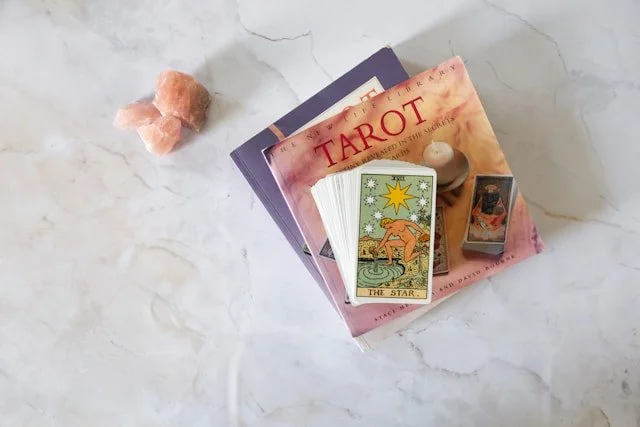 A stack of tarot books and cards on a white marble surface, with a pink salt rock and a small statue.