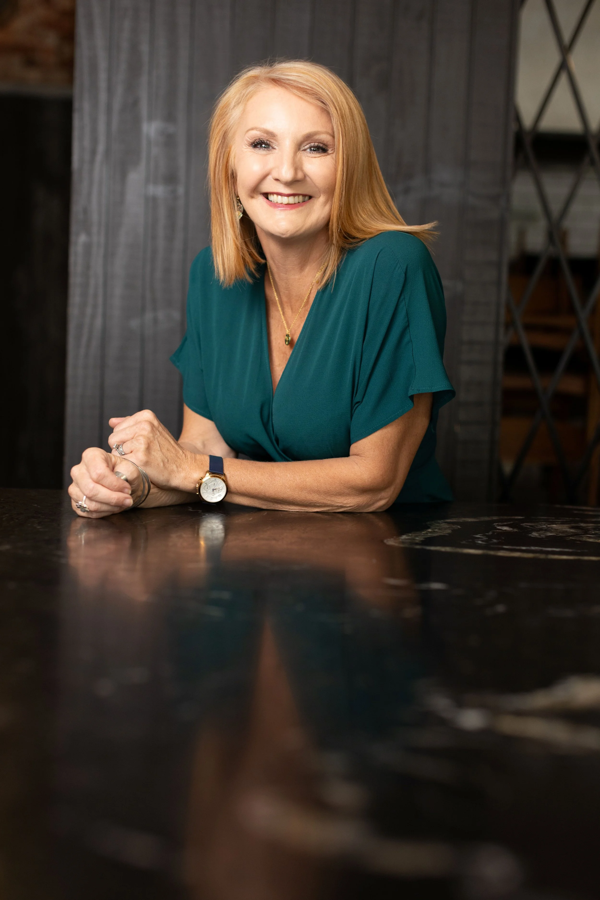 A smiling middle-aged woman with shoulder-length red hair, wearing a teal dress, gold jewelry, and a wristwatch, sitting at a dark wooden table indoors.