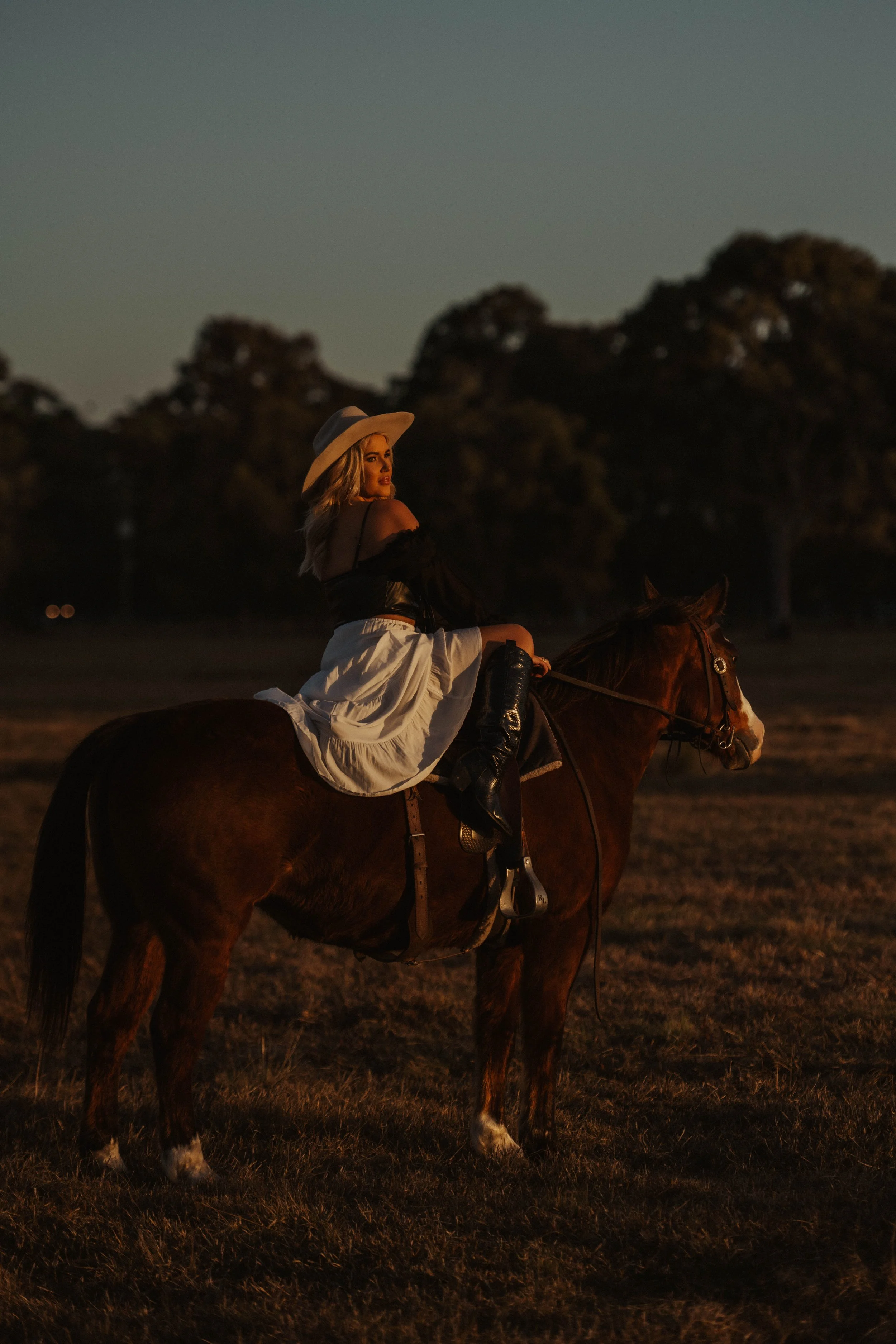 A woman riding a horse at dusk, wearing a wide-brimmed hat, black off-shoulder top, and a white skirt.