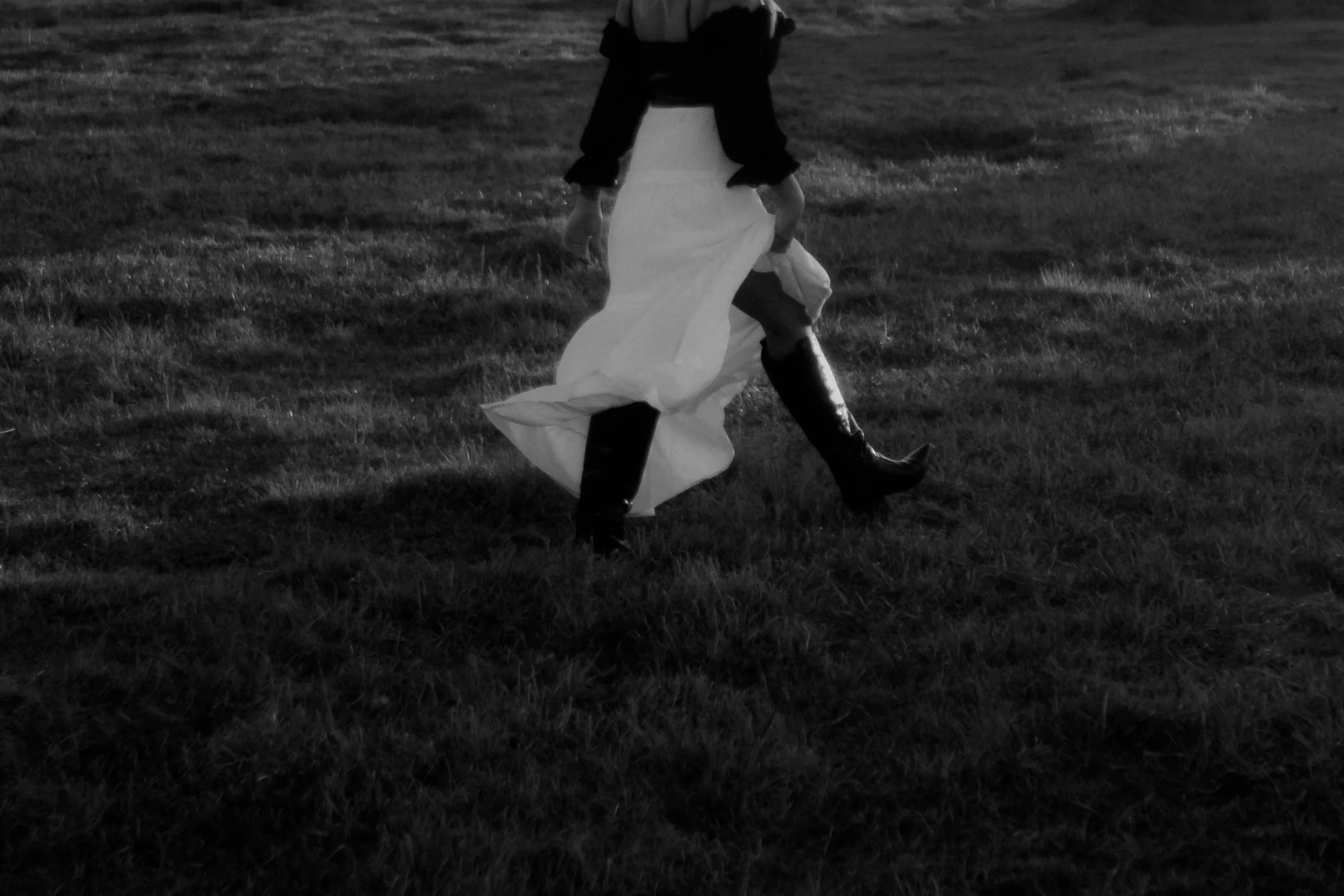 A person walking on grass wearing a long skirt, black jacket, and tall boots, with only their lower body visible.
