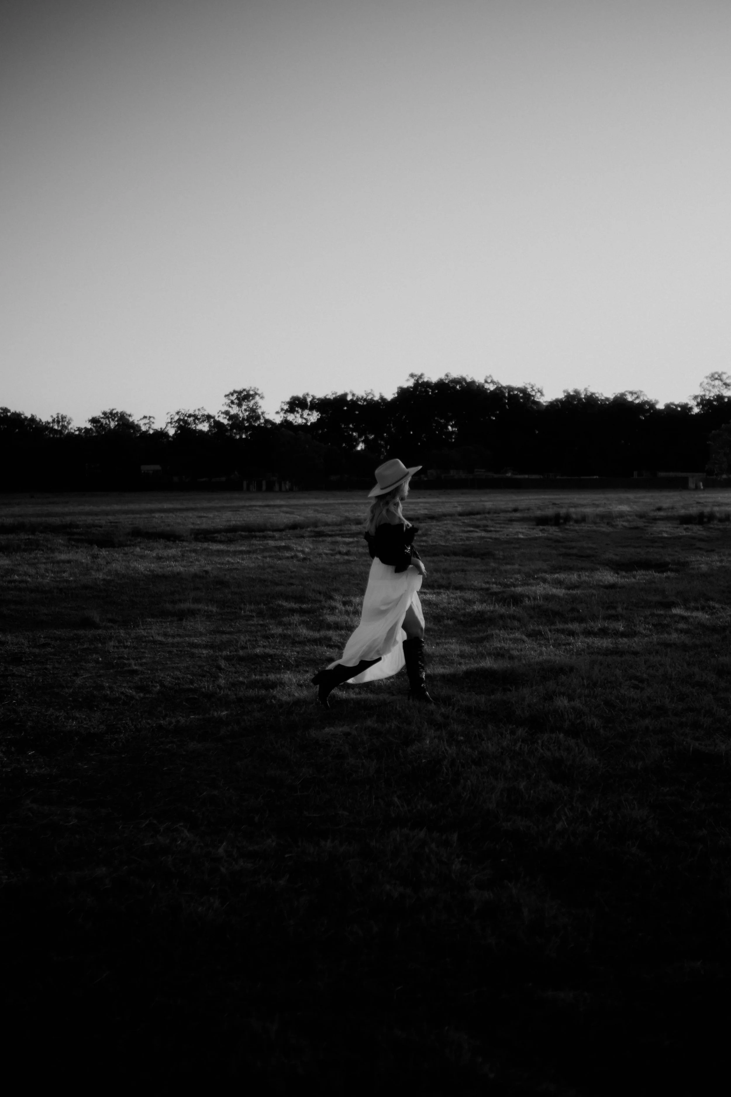 A woman wearing a wide-brimmed hat, black top, and long skirt walking across a grassy field during sunset, with trees in the background.