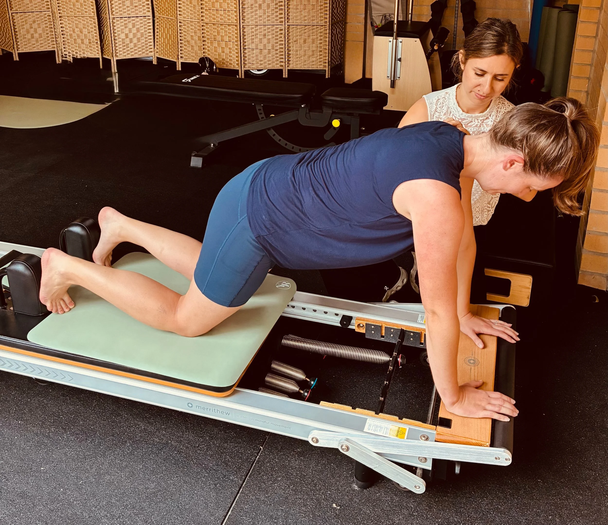 A woman performing a rehabilitation exercise on a Pilates reformer machine with a trainer guiding her.
