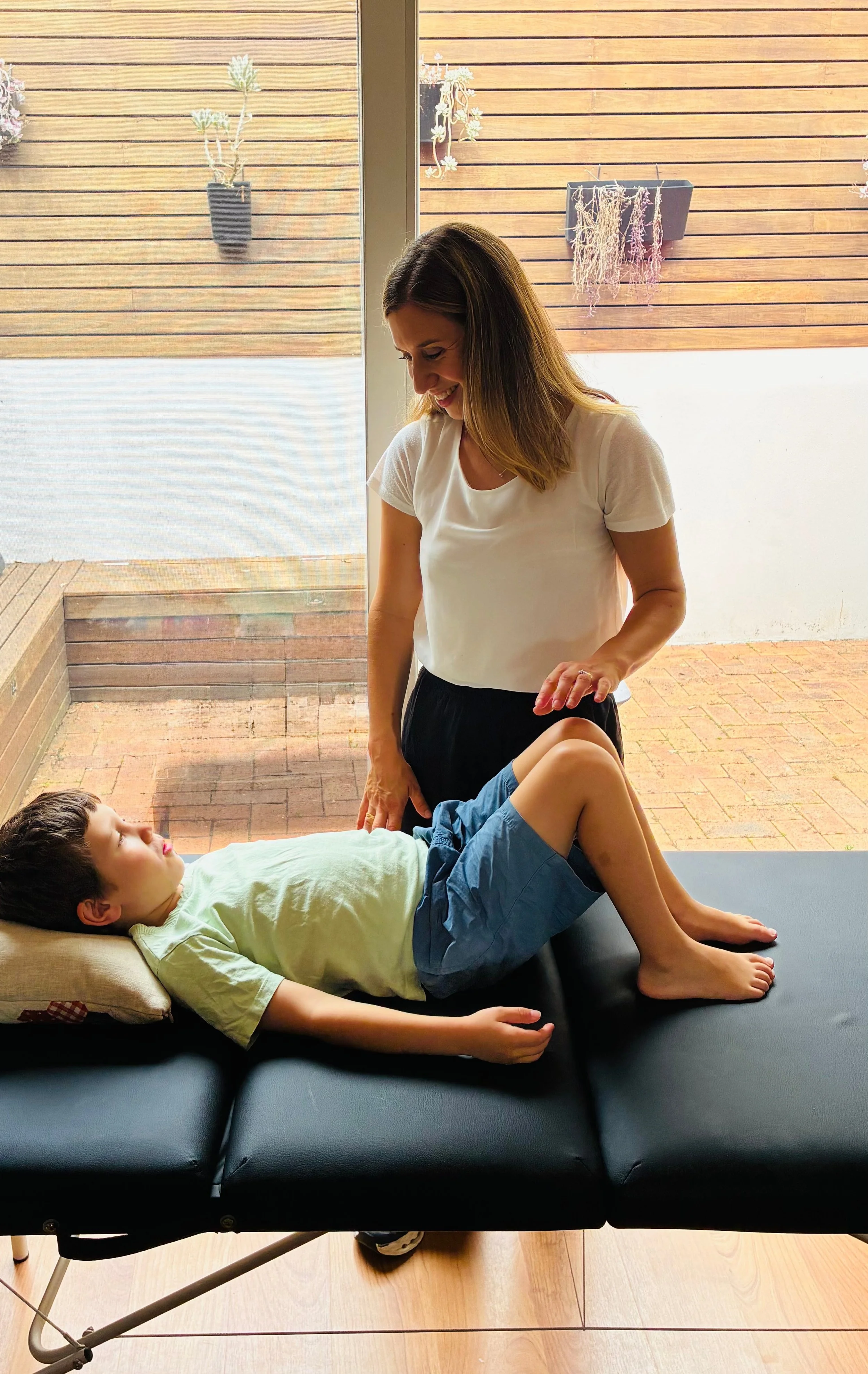 A woman practicing physical therapy on a young boy lying on a black therapy table indoors with a glass door and wooden deck outside.
