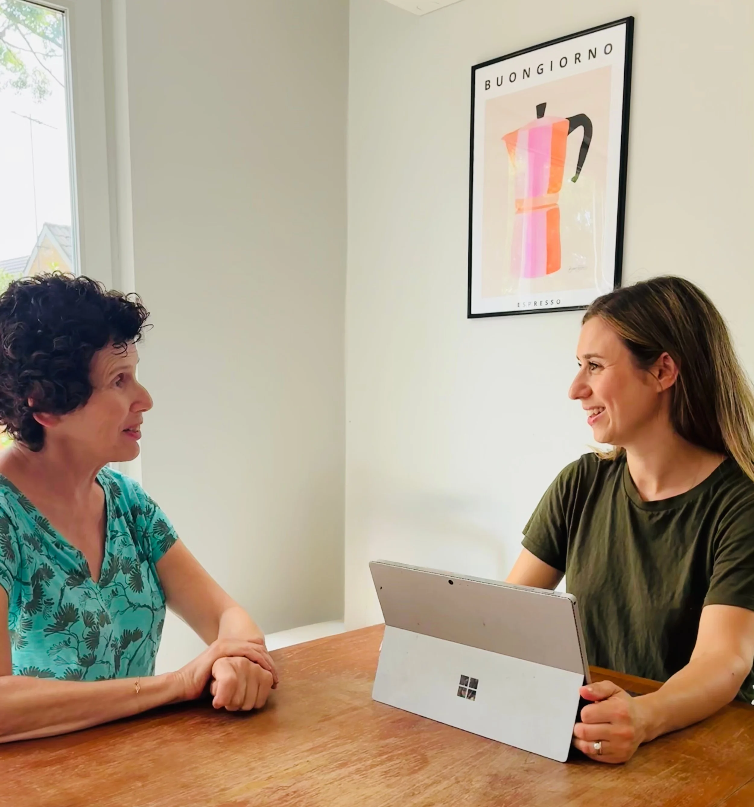 Two women sitting at a wooden table, having a conversation. The younger woman on the right uses a tablet and is smiling, while the older woman on the left listens attentively. A window is in the background, and a framed coffee-themed poster hangs on the wall.