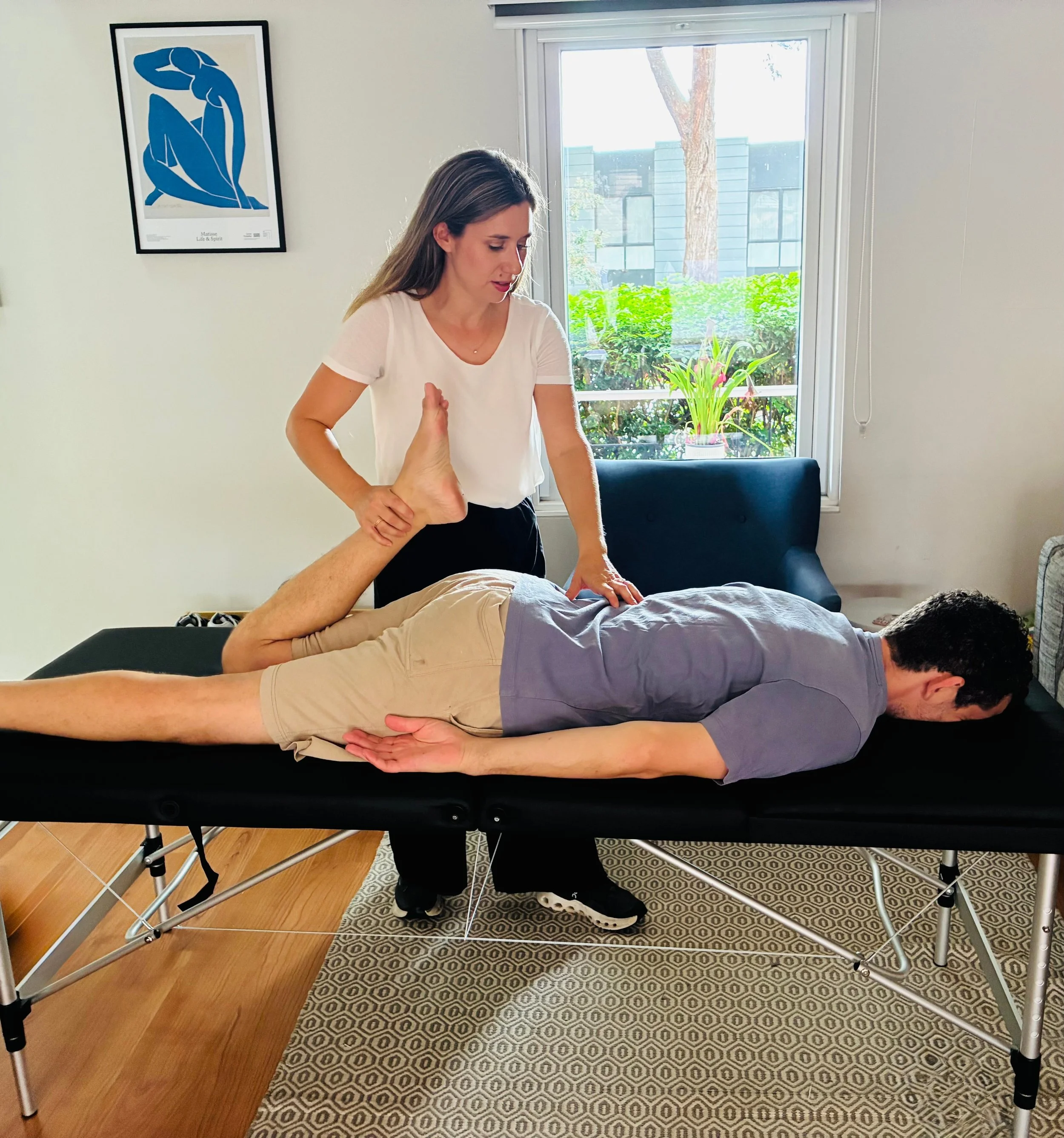 A woman is performing a medical or therapeutic treatment on a man lying face down on a massage table. The woman appears to be applying pressure or a massage technique on the man's side while holding his leg. The setting is a bright room with a window showing outdoor greenery.