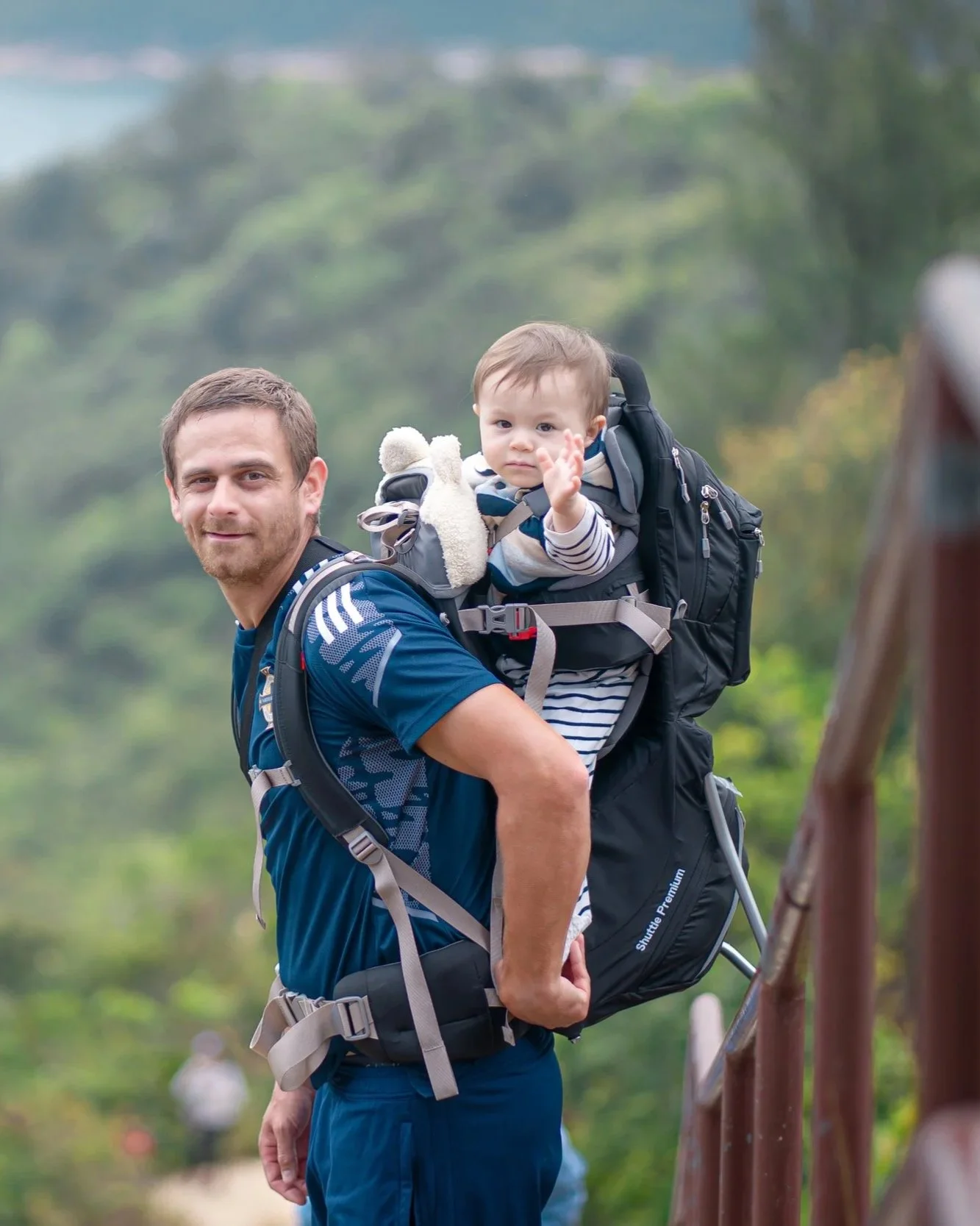 A man carrying a young child on his back outdoors in a green, wooded area.