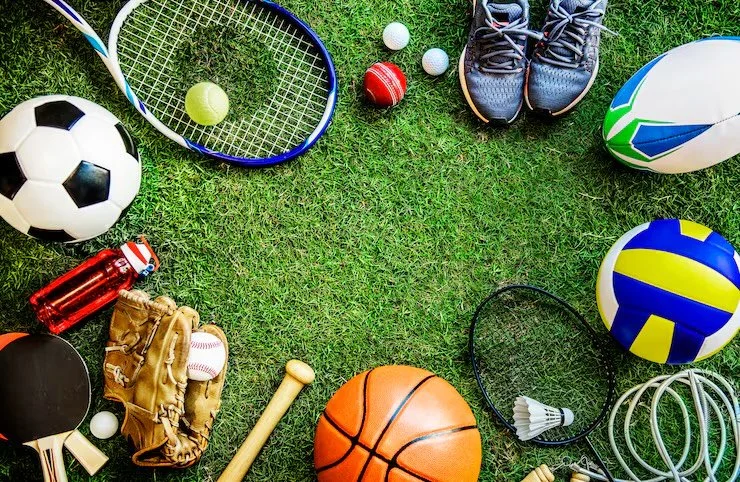 Various sports equipment arranged on grass, including a soccer ball, tennis racket and ball, golf clubs, baseball gloves and ball, basketball, volleyball, rugby ball, badminton racket, shuttlecock, water bottles, and tennis shoes.